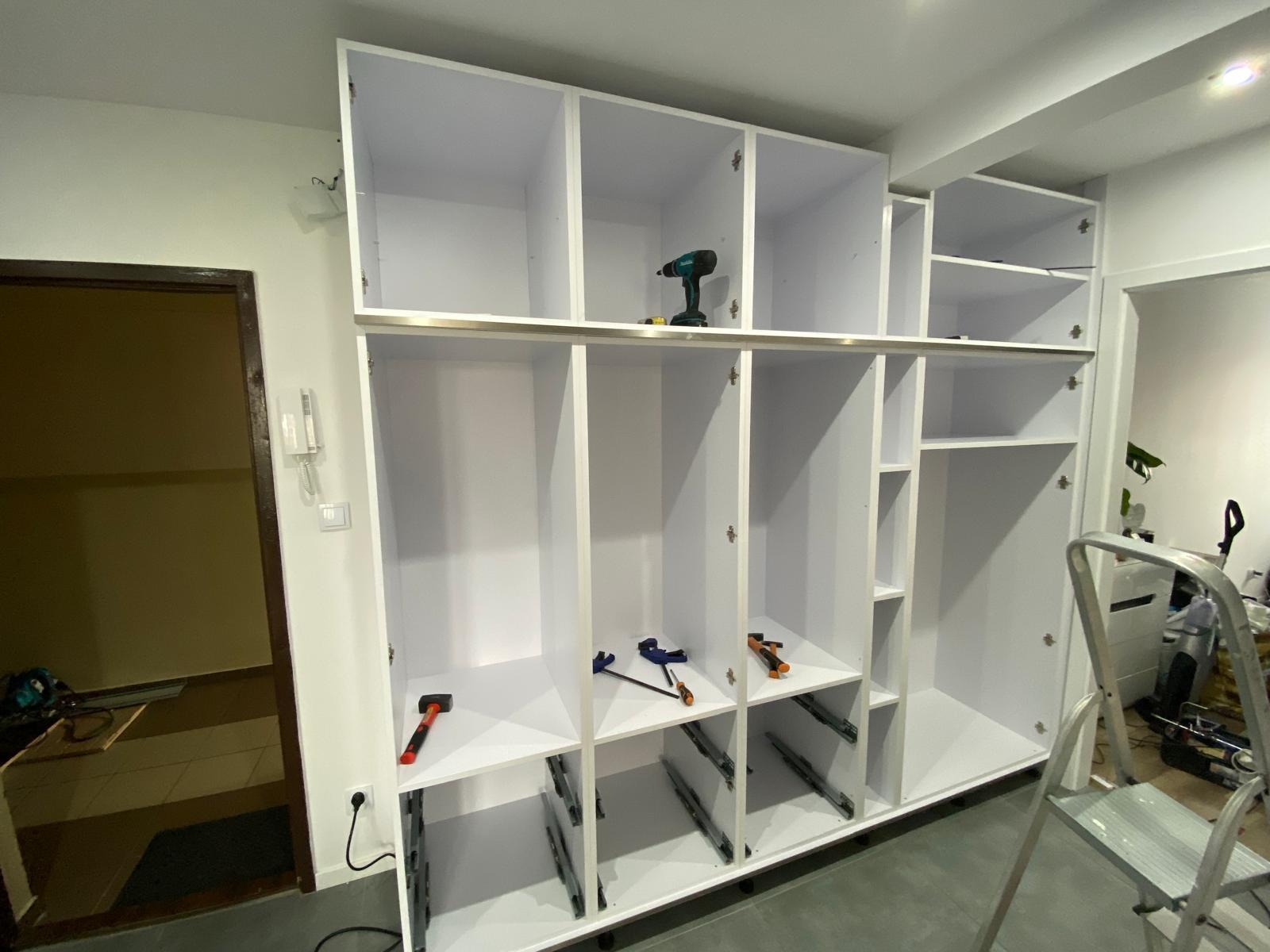 Wnętrze w trakcie montażu białej szafy wnękowej z narzędziami: wiertarka, młotek i ściski stolarskie. Widoczne szuflady z prowadnicami i elementy konstrukcyjne.