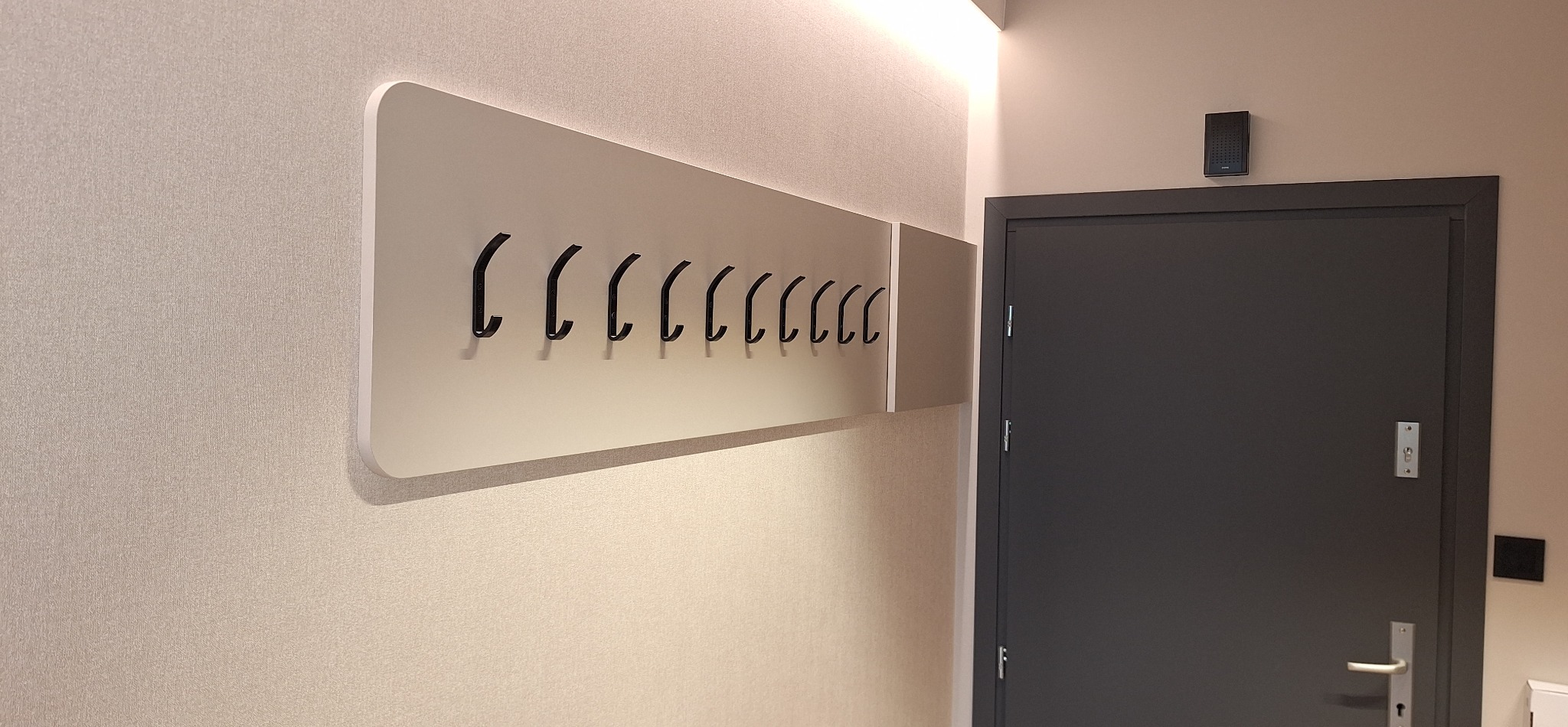 Nowoczesny wieszak ścienny z czarnymi haczykami na jasnej płycie, zamontowany w przedpokoju obok szarych drzwi z domofonem i oświetleniem LED na suficie.