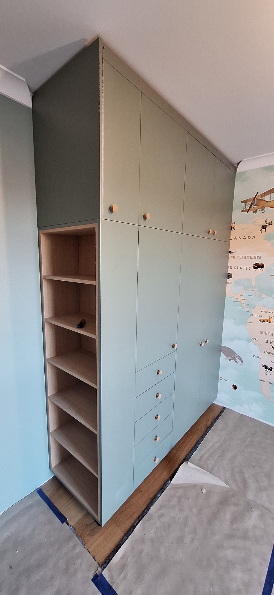 Miętowa szafa wnękowa z drewnianymi uchwytami, widoczna otwarta półka z narzędziem w środku, na ścianie mapa świata. Podłoga zabezpieczona folią malarską.