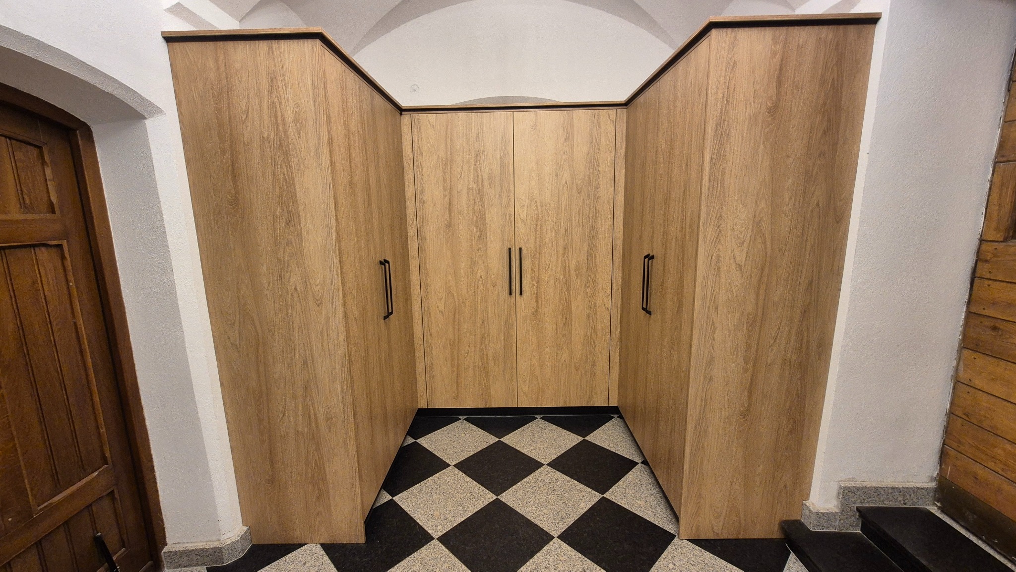 Funkcjonalna szafa wnękowa z drewnianymi frontami, idealnie dopasowana do nietypowej przestrzeni z czarno-białą podłogą w szachownicę i łukowym sufitem.