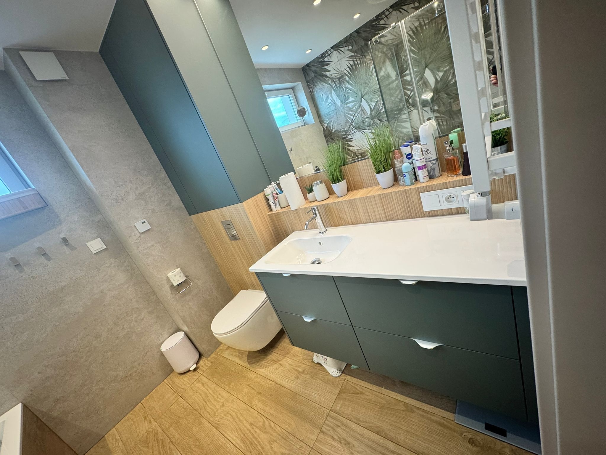 Nowoczesna łazienka z szafką pod umywalką w odcieniach zieleni, drewnianą podłogą i ścianą w szarości. Uwagę przykuwa lustro z roślinnym motywem w tle.