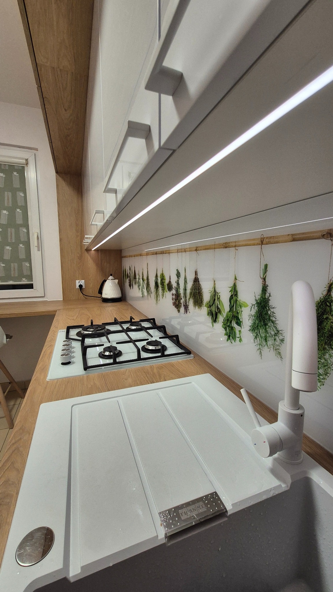 Jasna kuchnia z drewnianym blatem, białym zlewem Franke i płytą gazową. Nad blatem panel z motywem ziół i białe szafki podświetlone LED. Nowoczesny design.