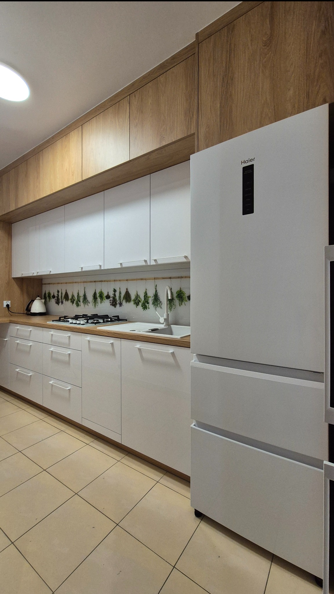 Nowoczesna kuchnia z białymi frontami i drewnianymi szafkami górnymi. Na blacie suszone zioła, obok biała lodówka Haier. Jasna podłoga z płytek.