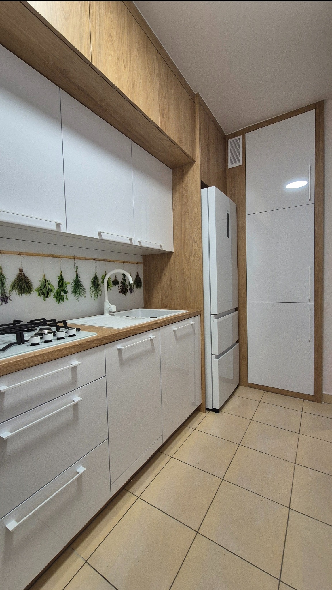 Nowoczesna kuchnia z białymi frontami i drewnianymi akcentami. Widoczny zlew, płyta gazowa i lodówka. Na ścianie dekoracja z suszonych ziół.
