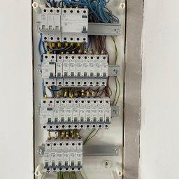 Inspektor Iskra - Rozdzielnica elektryczna Siemens z licznymi bezpiecznikami i okablowaniem, zamontowana w ścianie w trakcie wykończenia. Widoczne przewody w kolorach niebieskim, brązowym i żółto-zielonym.