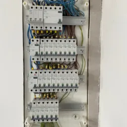 Rozdzielnica elektryczna Siemens z licznymi bezpiecznikami i okablowaniem, zamontowana w ścianie w trakcie wykończenia. Widoczne przewody w kolorach niebieskim, brązowym i żółto-zielonym.