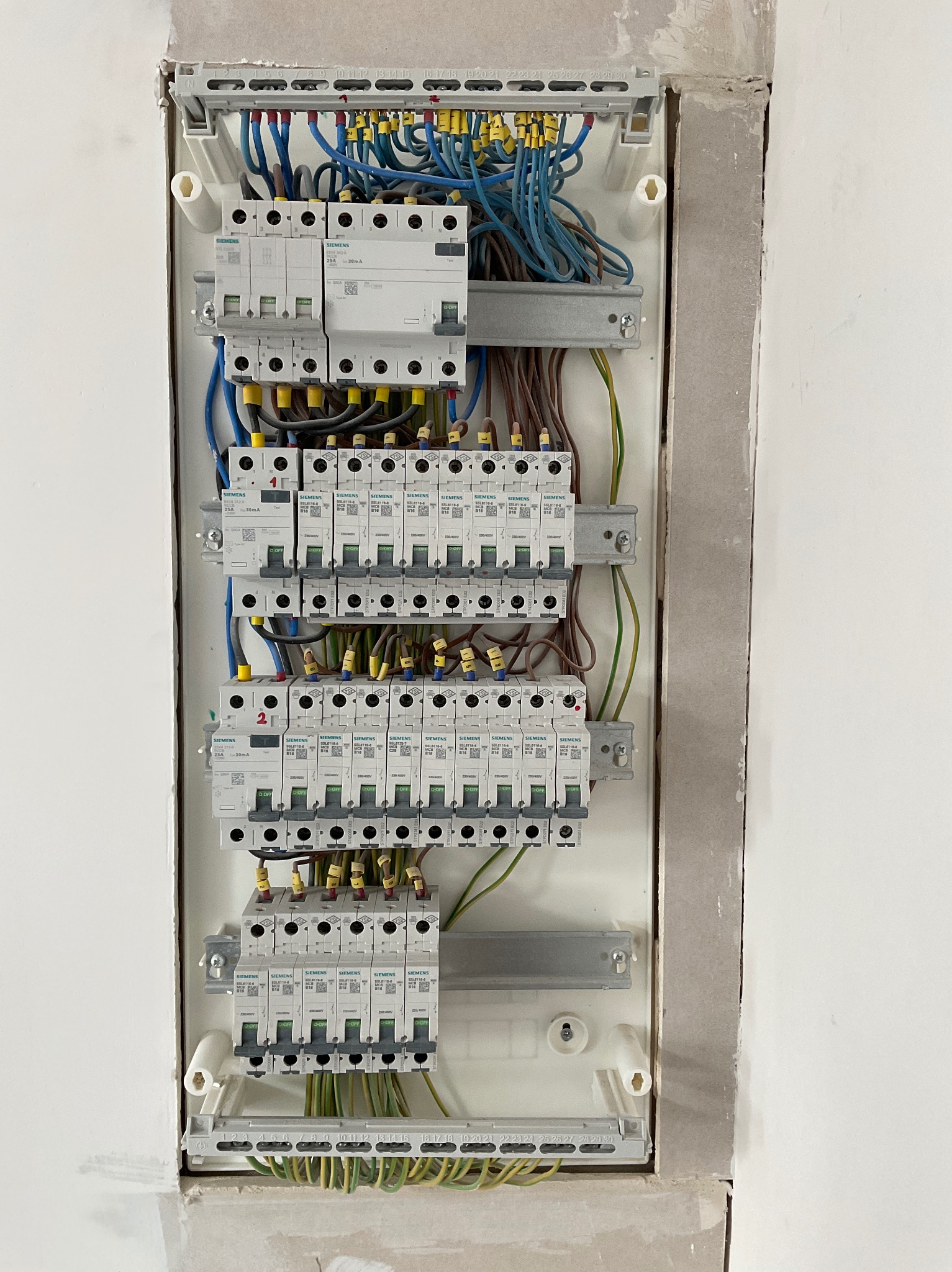 Rozdzielnica elektryczna Siemens z licznymi bezpiecznikami i okablowaniem, zamontowana w ścianie w trakcie wykończenia. Widoczne przewody w kolorach niebieskim, brązowym i żółto-zielonym.