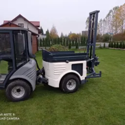 Mini wiertnica na trawniku, gotowa do pracy przy studni. Widoczny mechanizm wiertniczy i skrzynia ładunkowa wypełniona ziemią. W tle dom i rząd tuj.