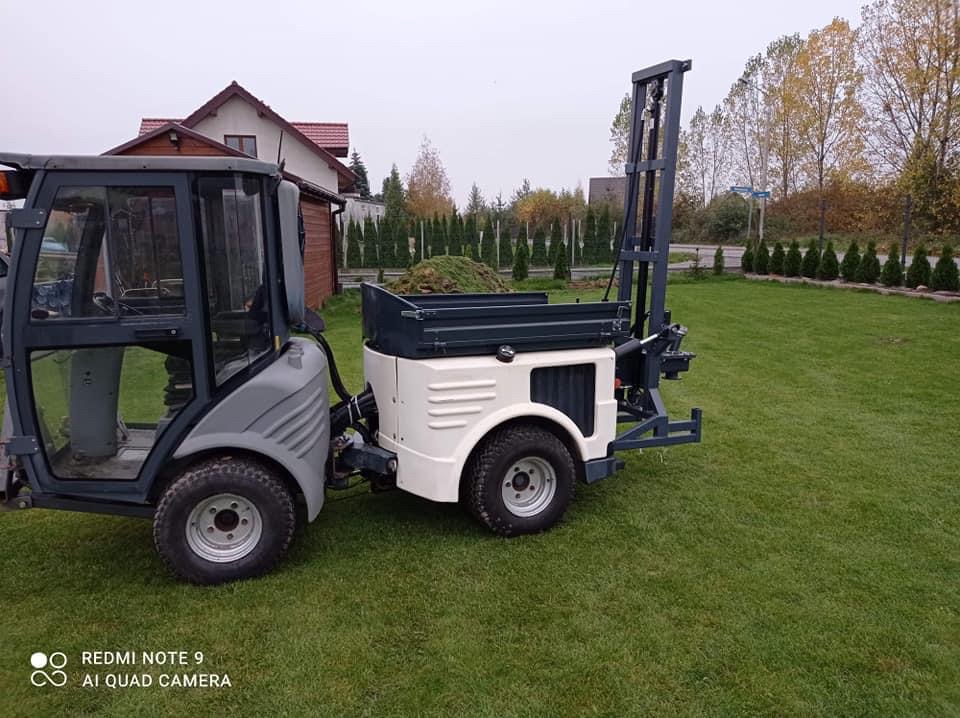 Mini wiertnica na trawniku, gotowa do pracy przy studni. Widoczny mechanizm wiertniczy i skrzynia ładunkowa wypełniona ziemią. W tle dom i rząd tuj.