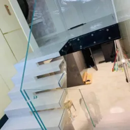 Nowoczesne schody z białego drewna z przezroczystą, szklaną balustradą, mocowaną punktowo. Widok z góry, oświetlenie sufitowe.