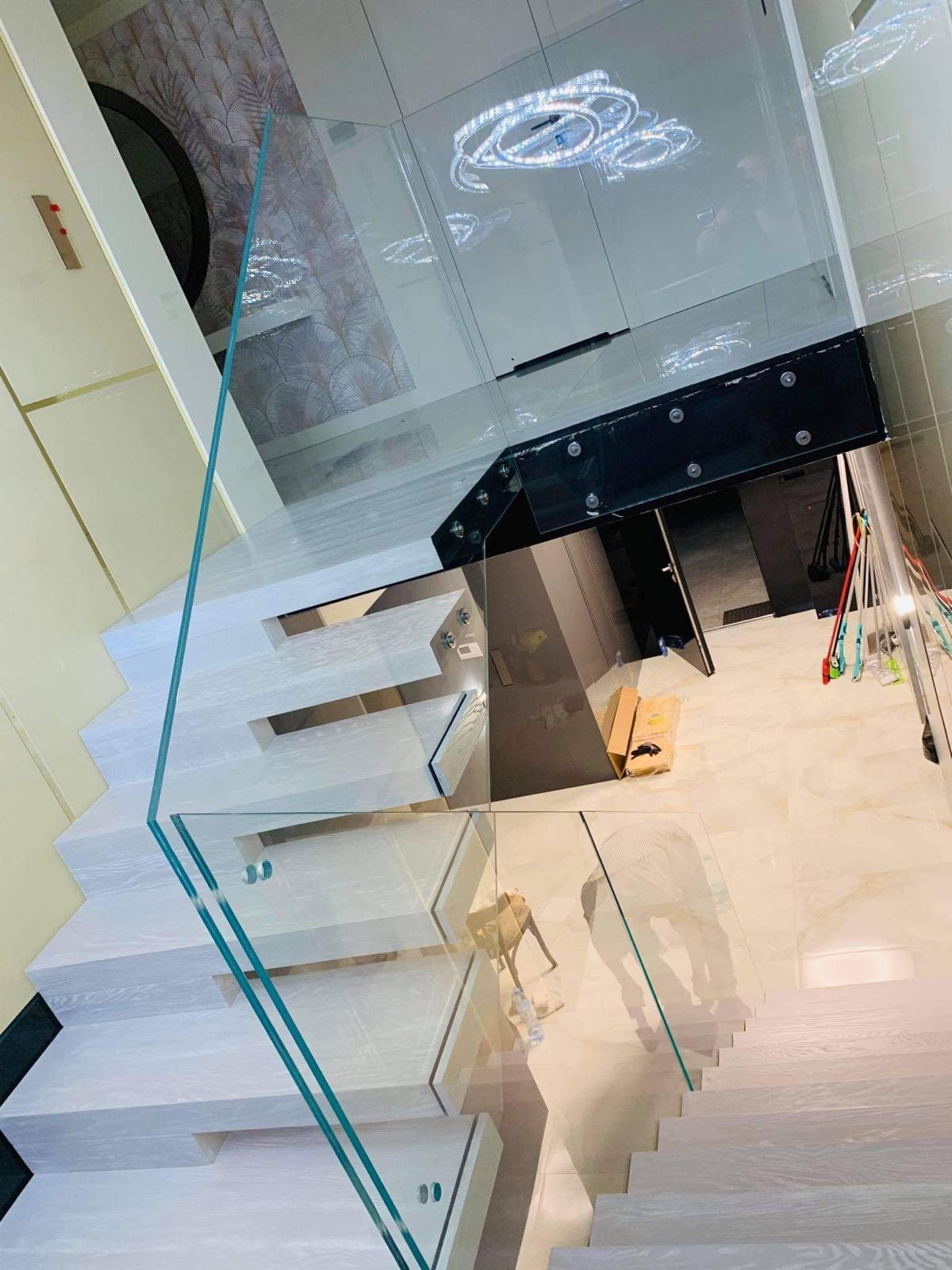 Nowoczesne schody z białego drewna z przezroczystą, szklaną balustradą, mocowaną punktowo. Widok z góry, oświetlenie sufitowe.