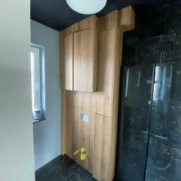 Nowoczesna łazienka z drewnianą zabudową nad stelażem WC, czarnym sufitem i marmurową kabiną prysznicową. Minimalistyczny design, oświetlenie sufitowe.