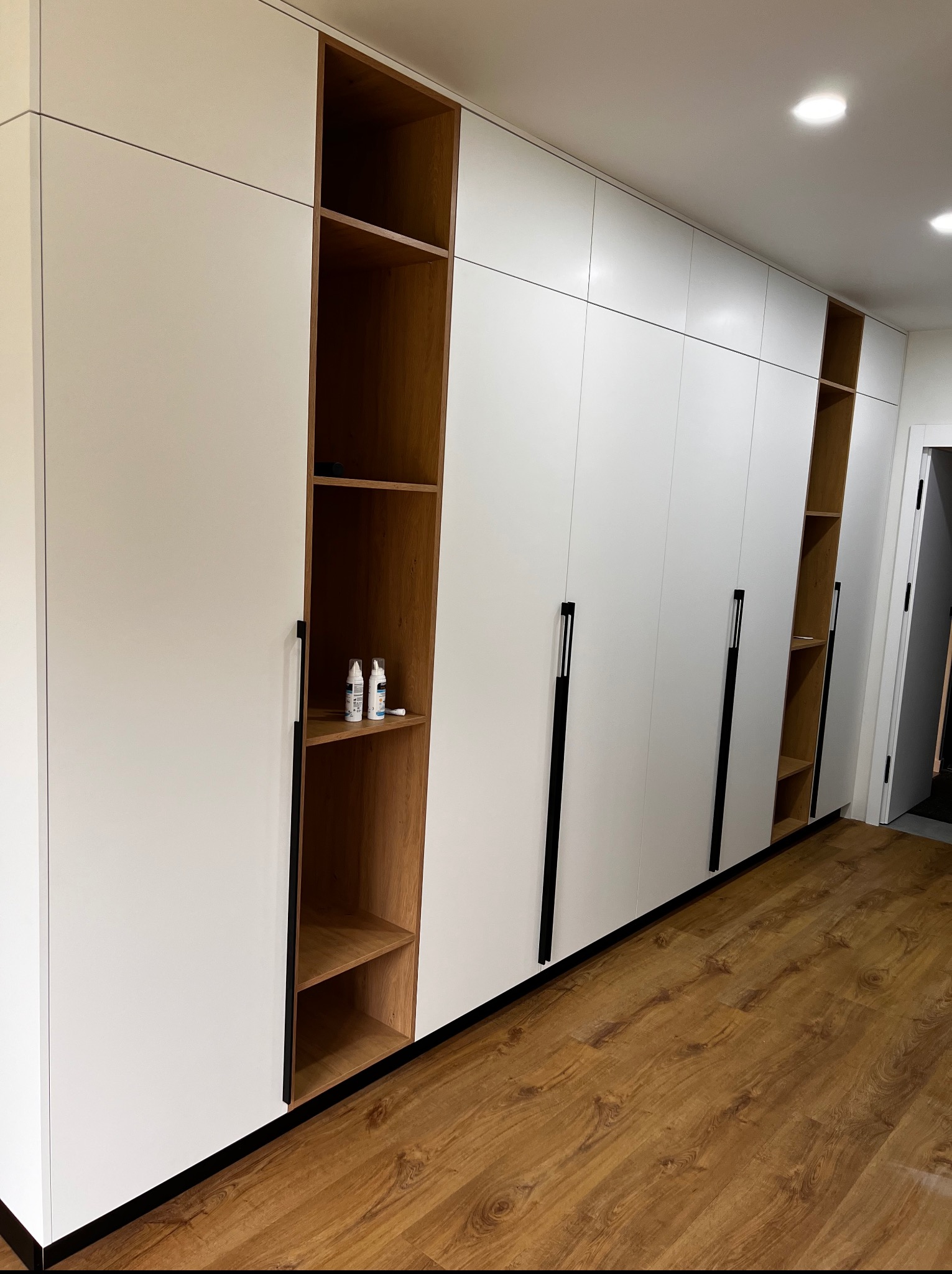 Biała szafa wnękowa z drewnianymi półkami i czarnymi uchwytami, na podłodze panele drewniane, oświetlenie sufitowe punktowe.