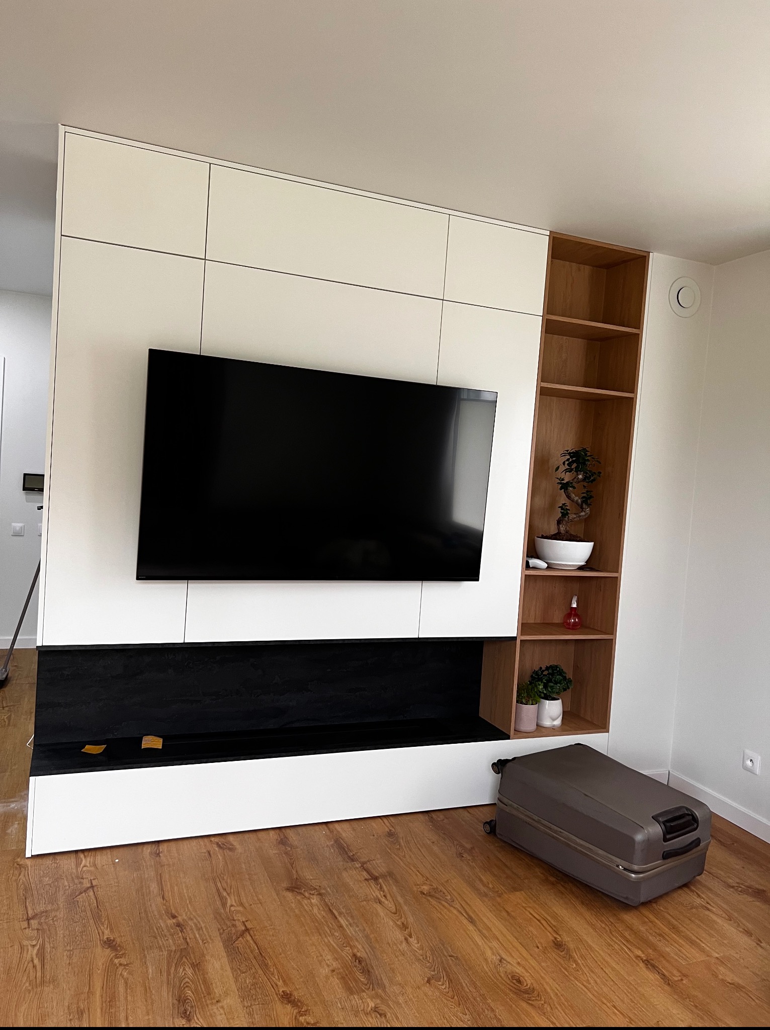 Biała szafa na wymiar z czarnym elementem dekoracyjnym pod telewizorem i drewnianym regałem z półkami po prawej stronie. Na podłodze walizka.