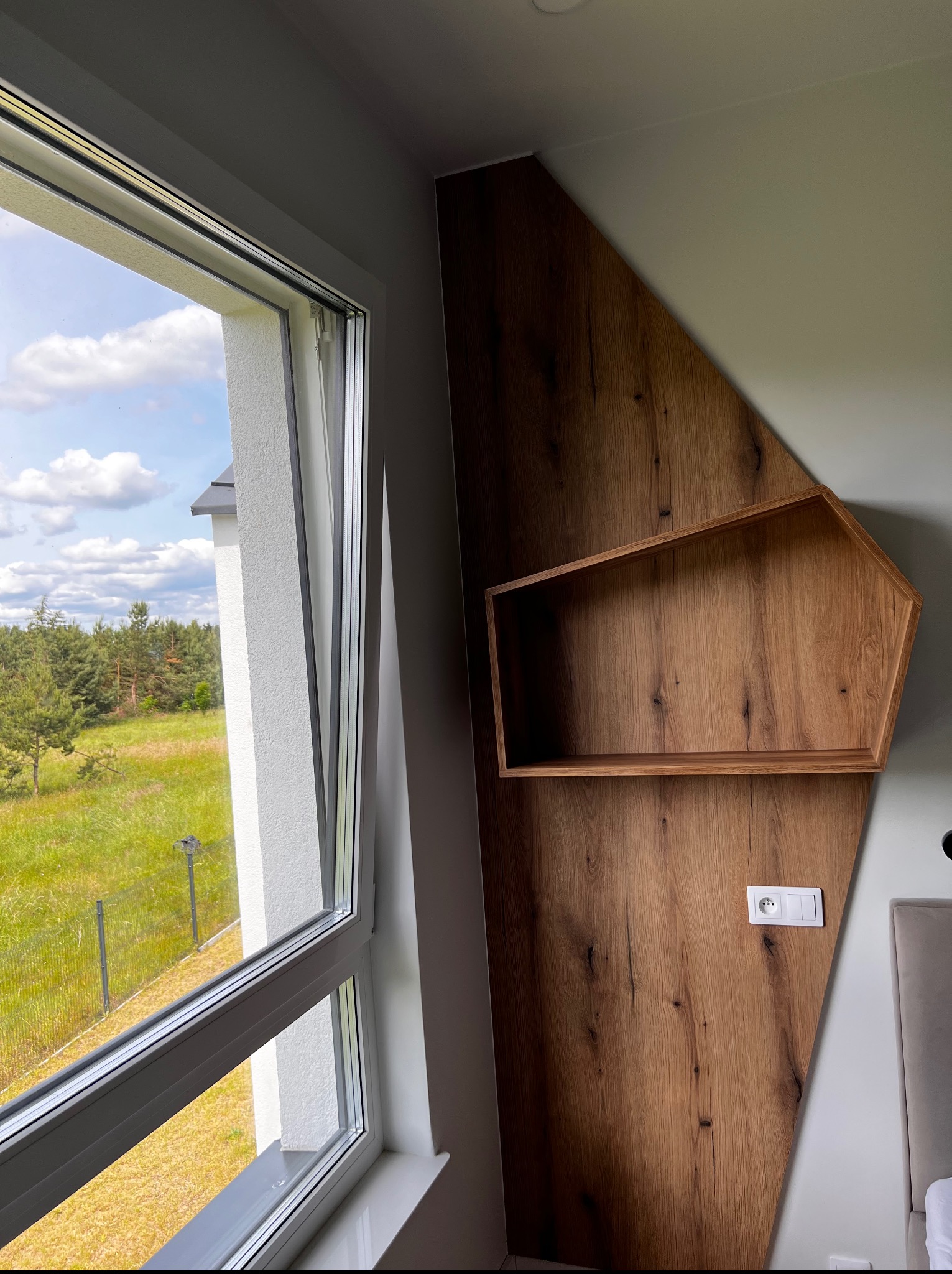Drewniana półka o nietypowym kształcie na ścianie z drewnianą okładziną, obok okna z widokiem na zieleń. Nowoczesne wnętrze, detale wykończenia, naturalne materiały.