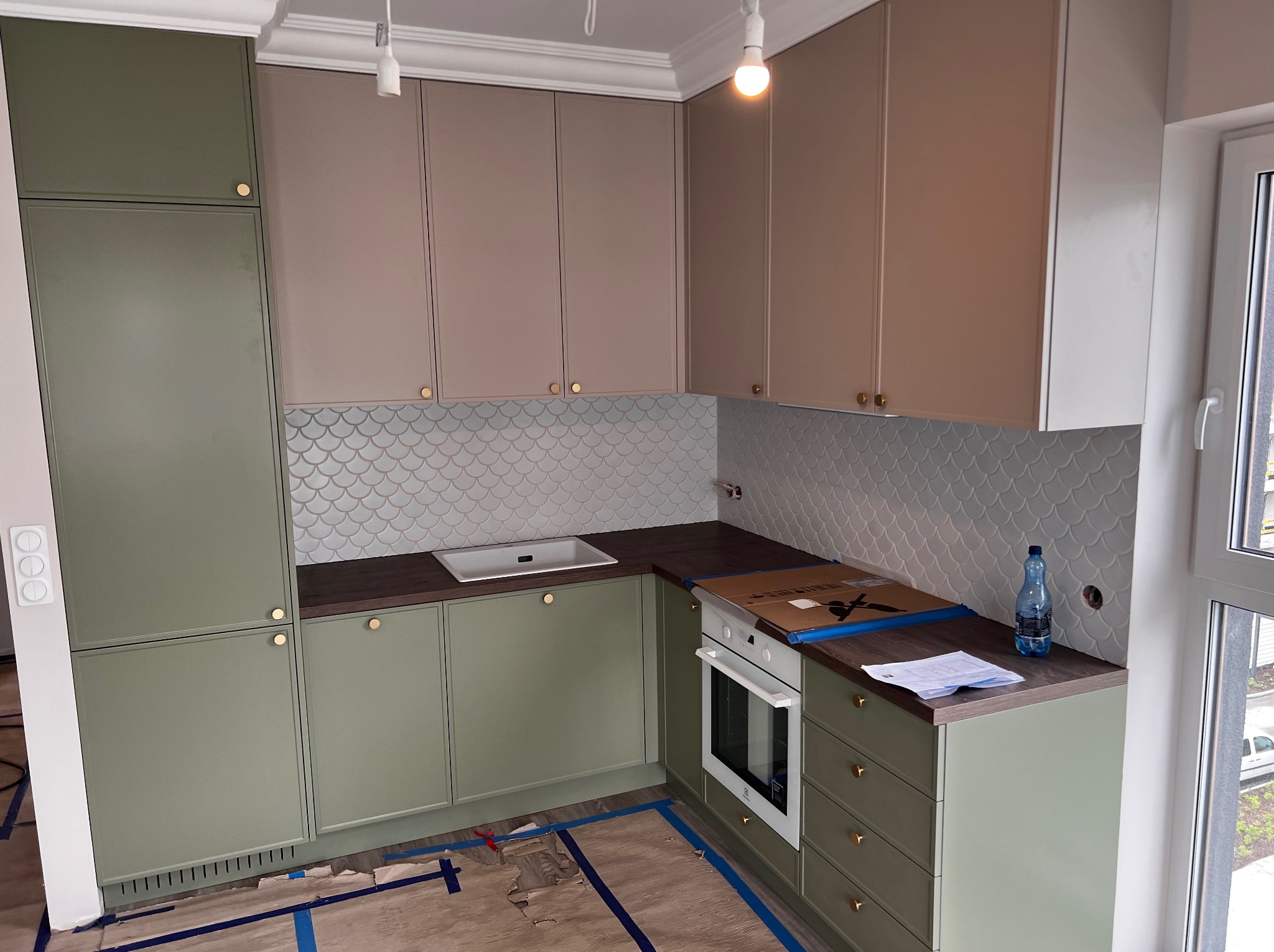 Kuchenne szafki na wymiar w odcieniach zieleni i beżu, z złotymi uchwytami. Na blacie widać dokumenty i butelkę wody. Tło stanowi mozaika w kształcie rybiej łuski.