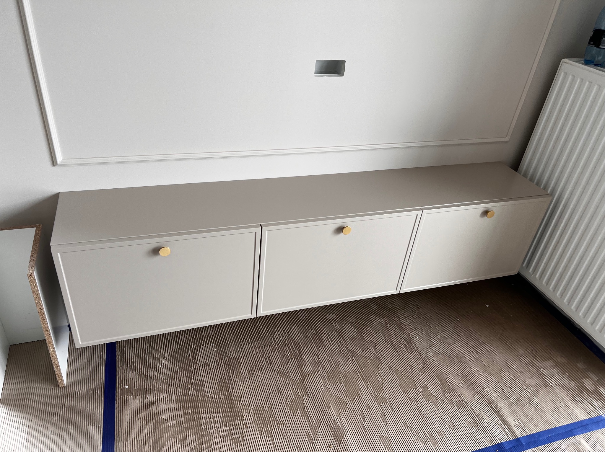 Jasnoszare, wiszące szafki z okrągłymi, złotymi uchwytami, zamontowane na ścianie z ozdobną listwą. Podłoga zabezpieczona folią malarską.