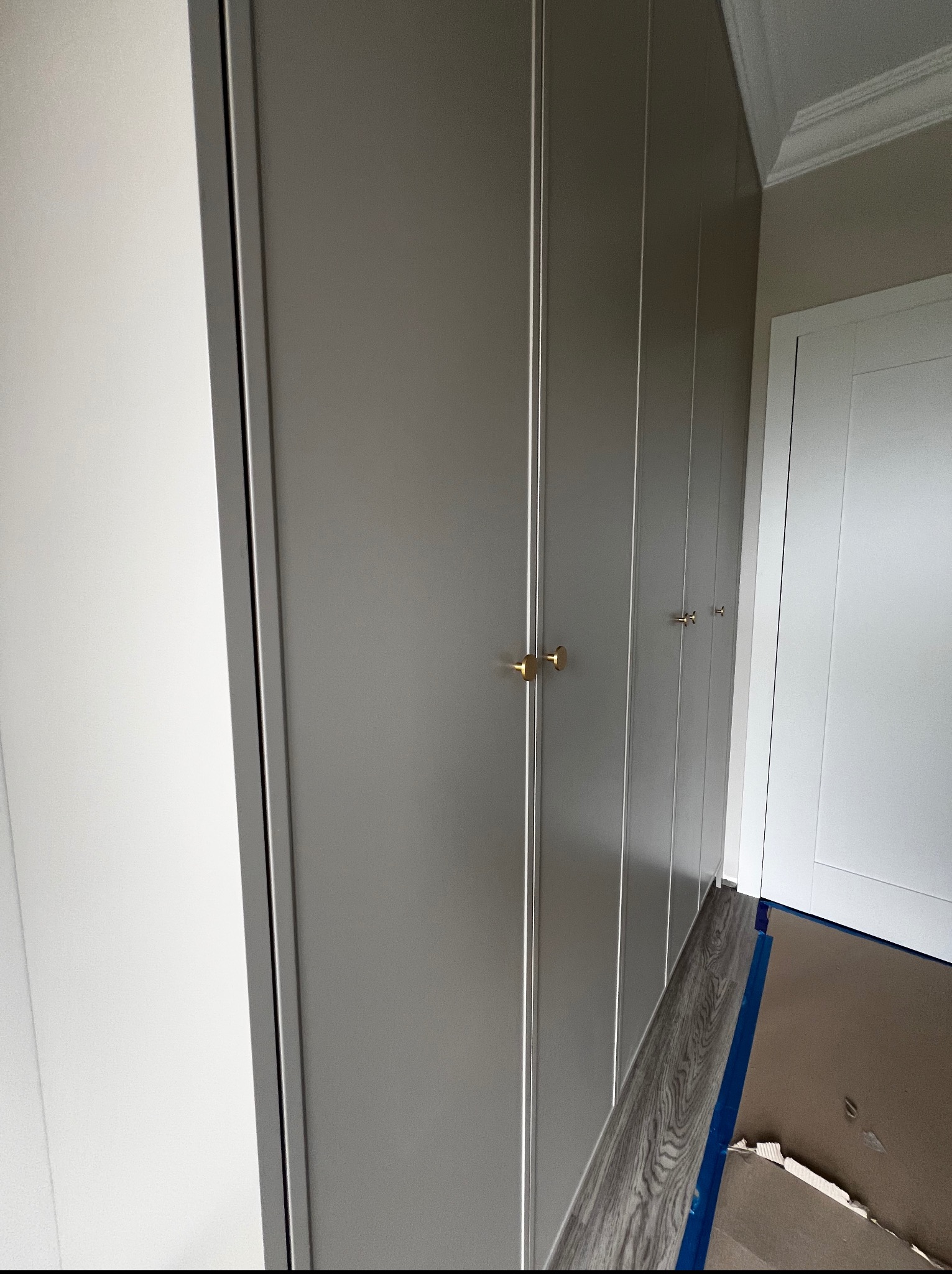 Szara szafa wnękowa z subtelnymi złotymi uchwytami, z widoczną listwą przypodłogową i fragmentem podłogi, w minimalistycznym wnętrzu.