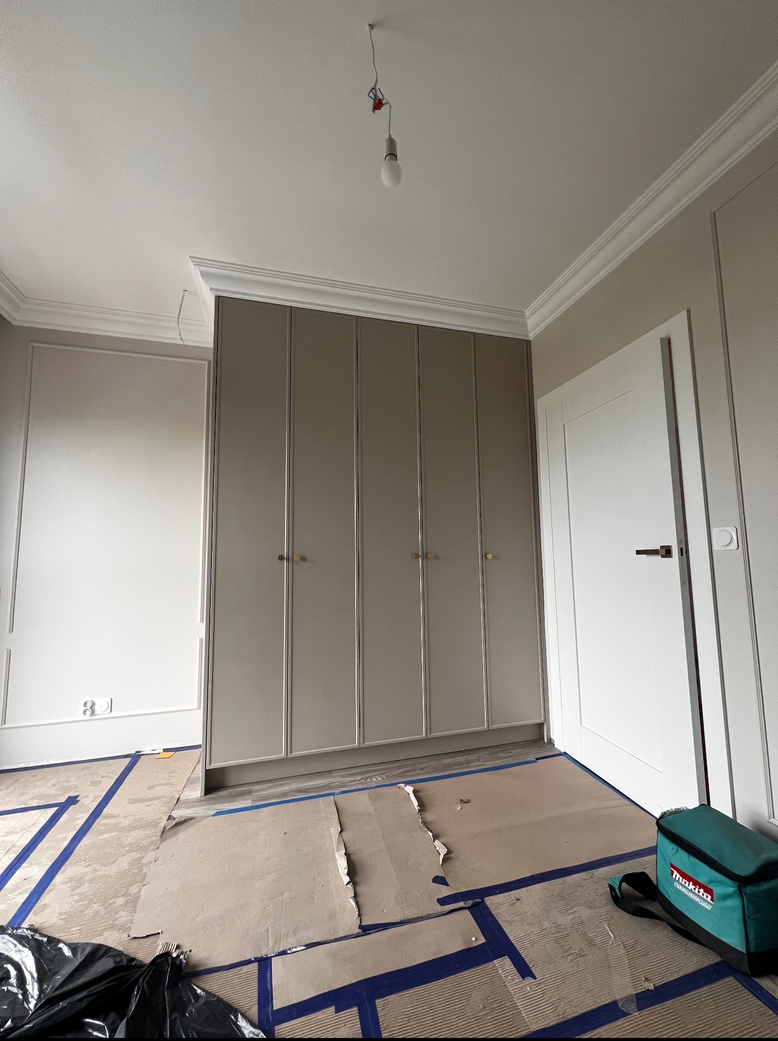 Beżowa szafa wnękowa z subtelnymi złotymi uchwytami, w pokoju z listwami dekoracyjnymi i zabezpieczoną podłogą, widok z dołu, w trakcie montażu.