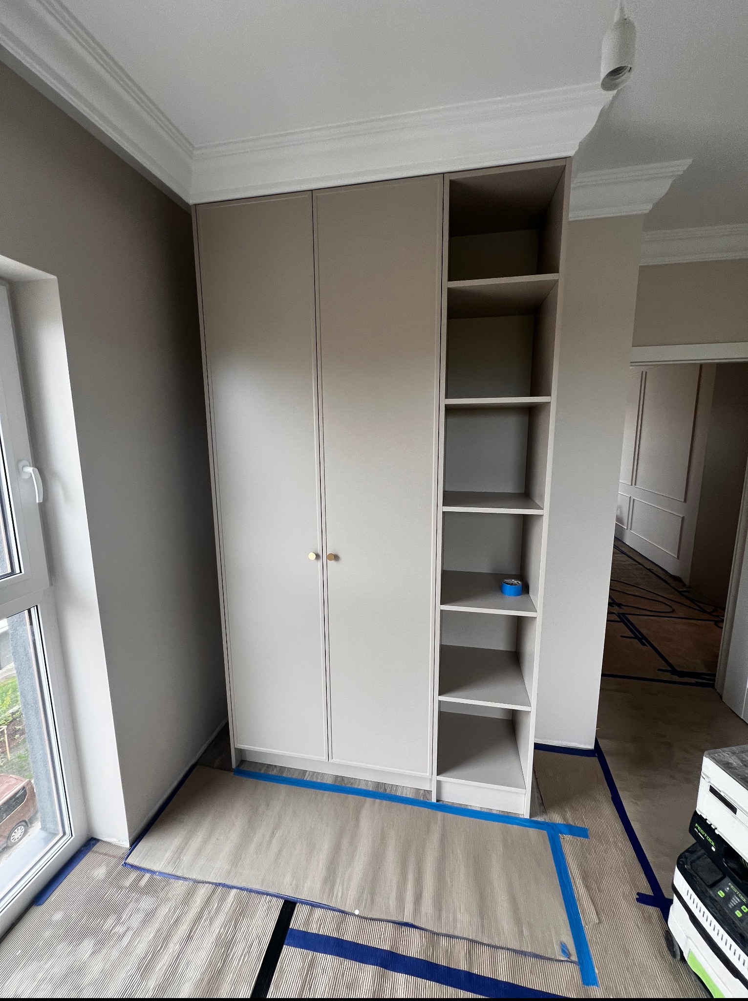 Jasna szafa na wymiar z otwartymi półkami, złote gałki, wykończenie listwami przysufitowymi. Podłoga zabezpieczona folią i taśmą malarską.