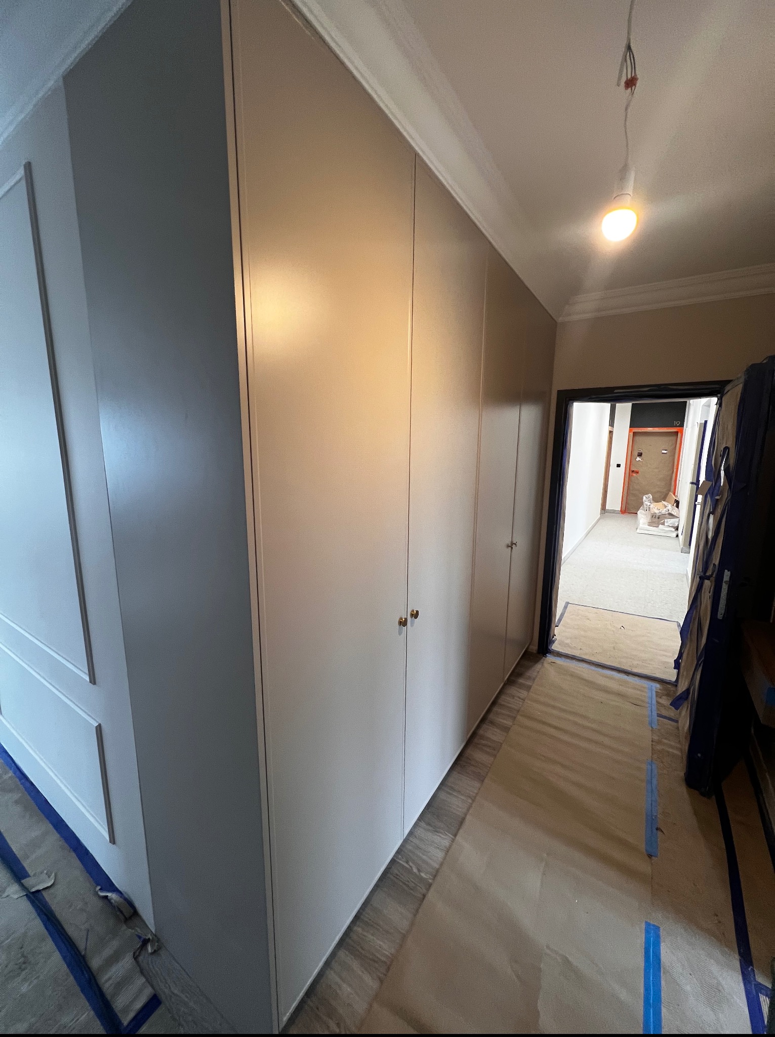 Długa, szara szafa wnękowa z złotymi uchwytami w korytarzu, zabezpieczona folią malarską. Widok na otwarte drzwi do dalszej części mieszkania.