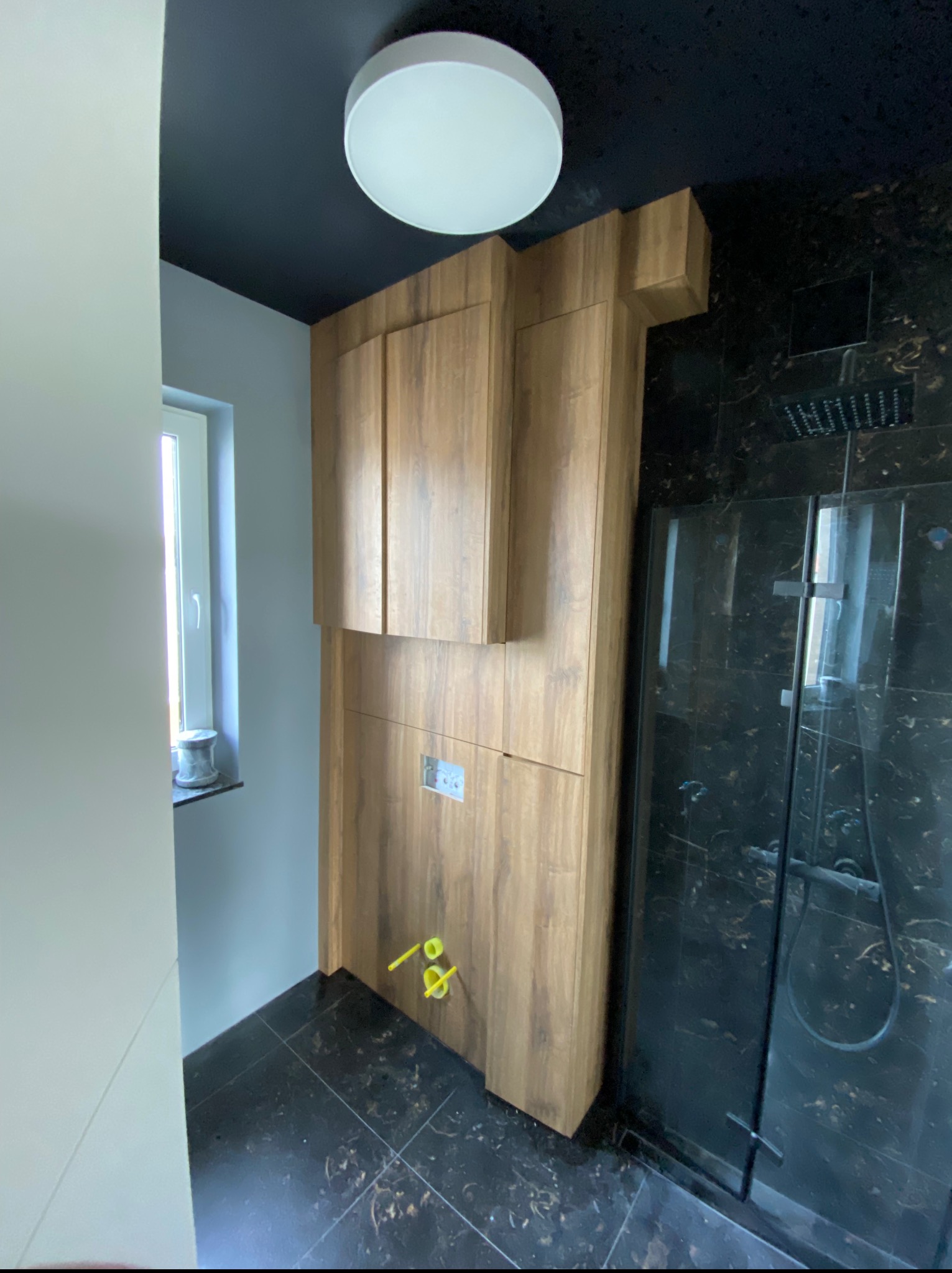 Nowoczesna łazienka z drewnianą zabudową nad stelażem WC, czarnym sufitem i marmurową kabiną prysznicową. Minimalistyczny design, oświetlenie sufitowe.