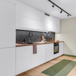 Michal Blaszczyk - Nowoczesna kuchnia z białymi szafkami, drewnianym blatem i czarnym panelem nad zlewem. Oświetlenie punktowe, roślina doniczkowa, dywanik. Minimalistyczny design.