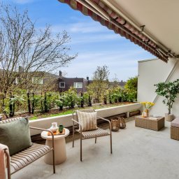 Michal Blaszczyk - Umeblowany balkon z markizą i widokiem na zieleń. Dwa krzesła, stolik i sofa tworzą przytulną przestrzeń. Rośliny wzdłuż balustrady dodają naturalnego akcentu.