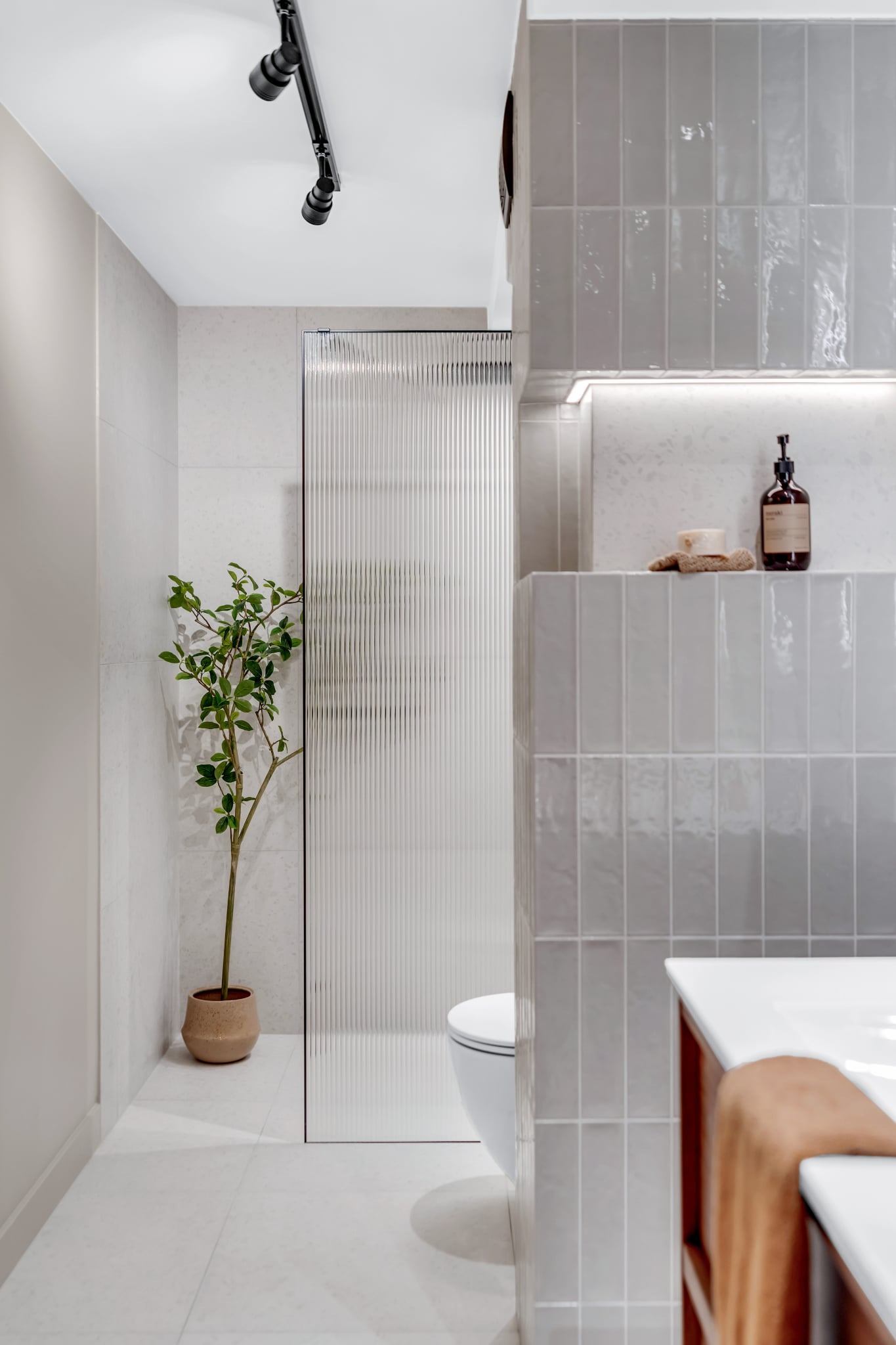 Jasna łazienka z szarymi, błyszczącymi płytkami ułożonymi pionowo, szklaną kabiną prysznicową i rośliną w doniczce. Minimalistyczny design, oświetlenie punktowe.