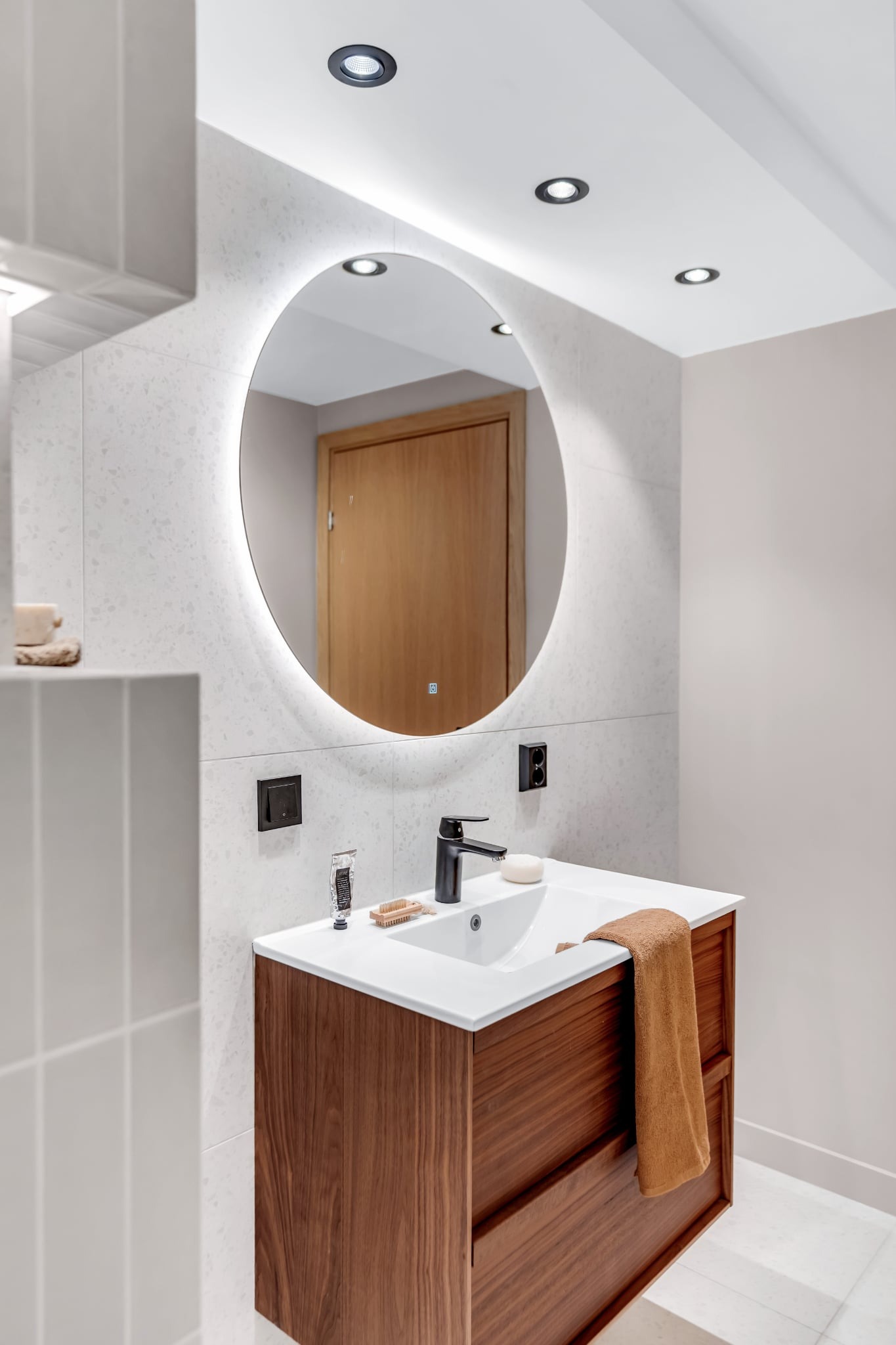 Nowoczesna łazienka z okrągłym lustrem LED, umywalką nablatową na szafce z drewna orzechowego i jasnymi płytkami imitującymi kamień na ścianie.
