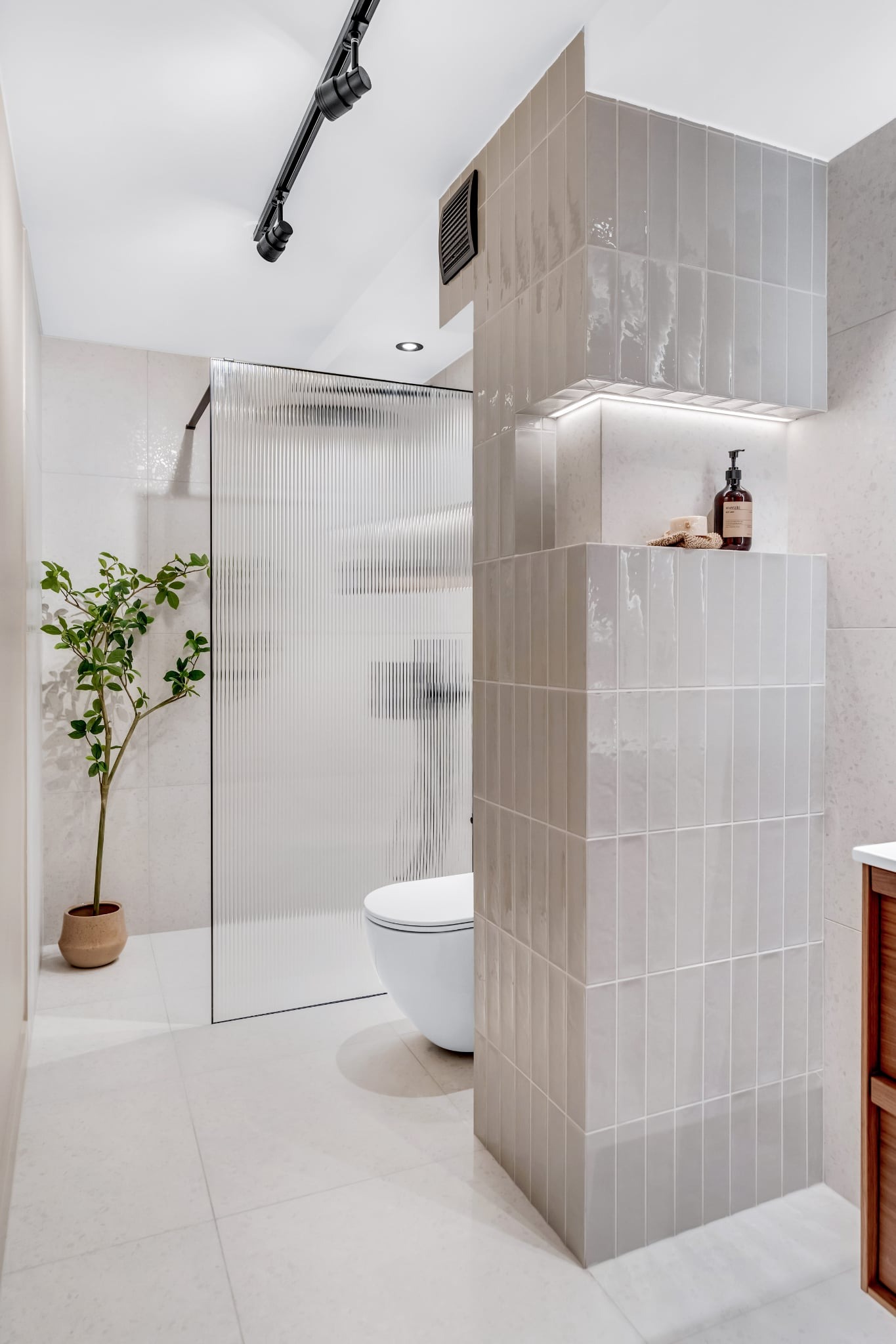 Nowoczesna łazienka z kabiną prysznicową z ryflowanego szkła, wiszącą toaletą i elementami wykończenia z płytek w odcieniach szarości. Minimalistyczny design z akcentem roślinnym.