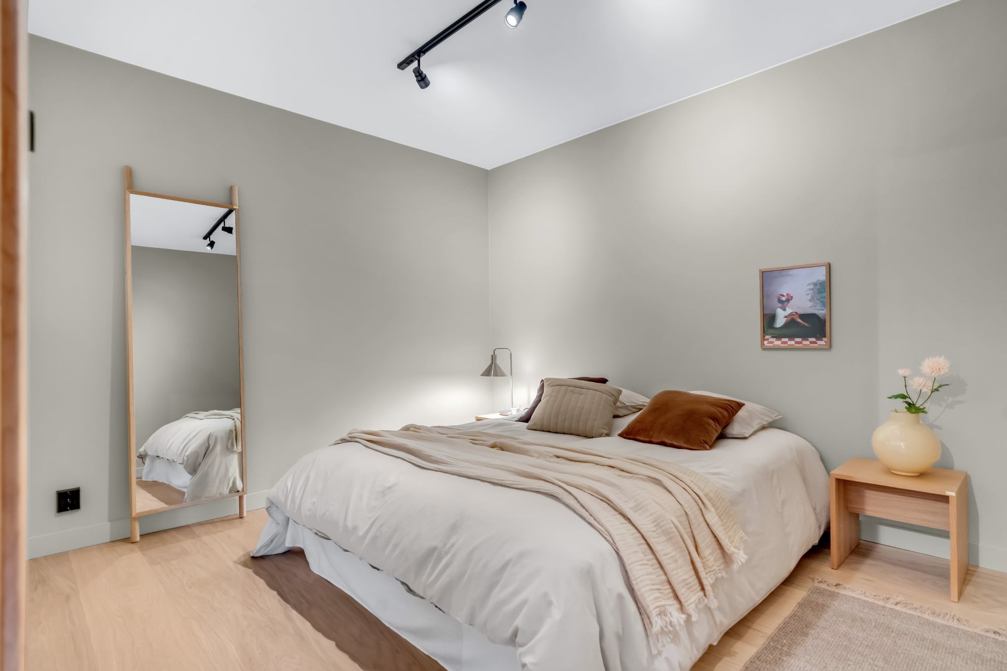 Neutralna sypialnia z drewnianą podłogą, łóżkiem z beżową pościelą, lustrem w drewnianej ramie i małym stolikiem nocnym z wazonem. Minimalistyczny design, stonowana kolorystyka.