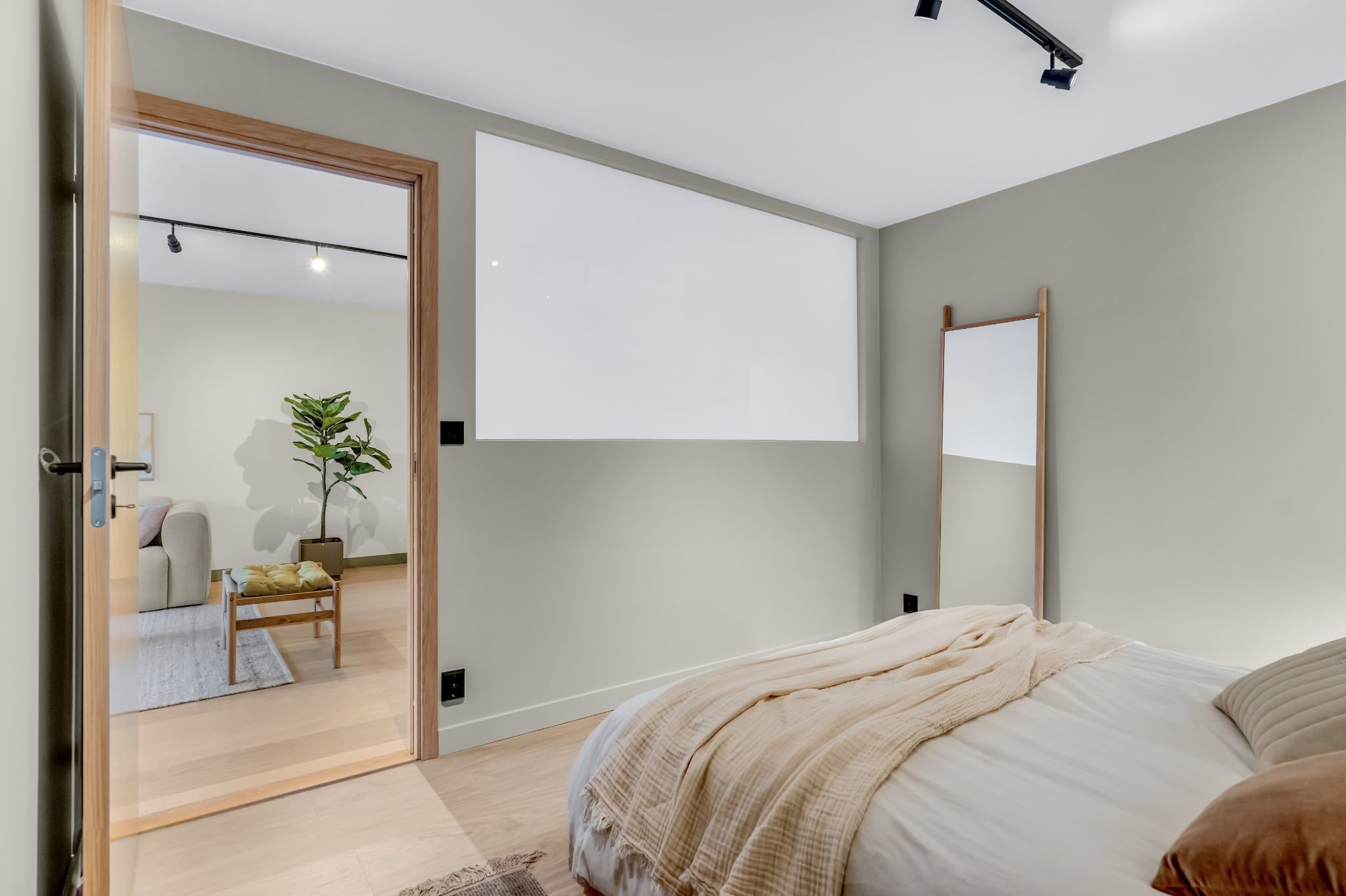 Nowoczesna sypialnia z beżową pościelą, dużym lustrem i widokiem na salon przez otwarte drzwi. Minimalistyczny design, stonowana kolorystyka, drewniane elementy.