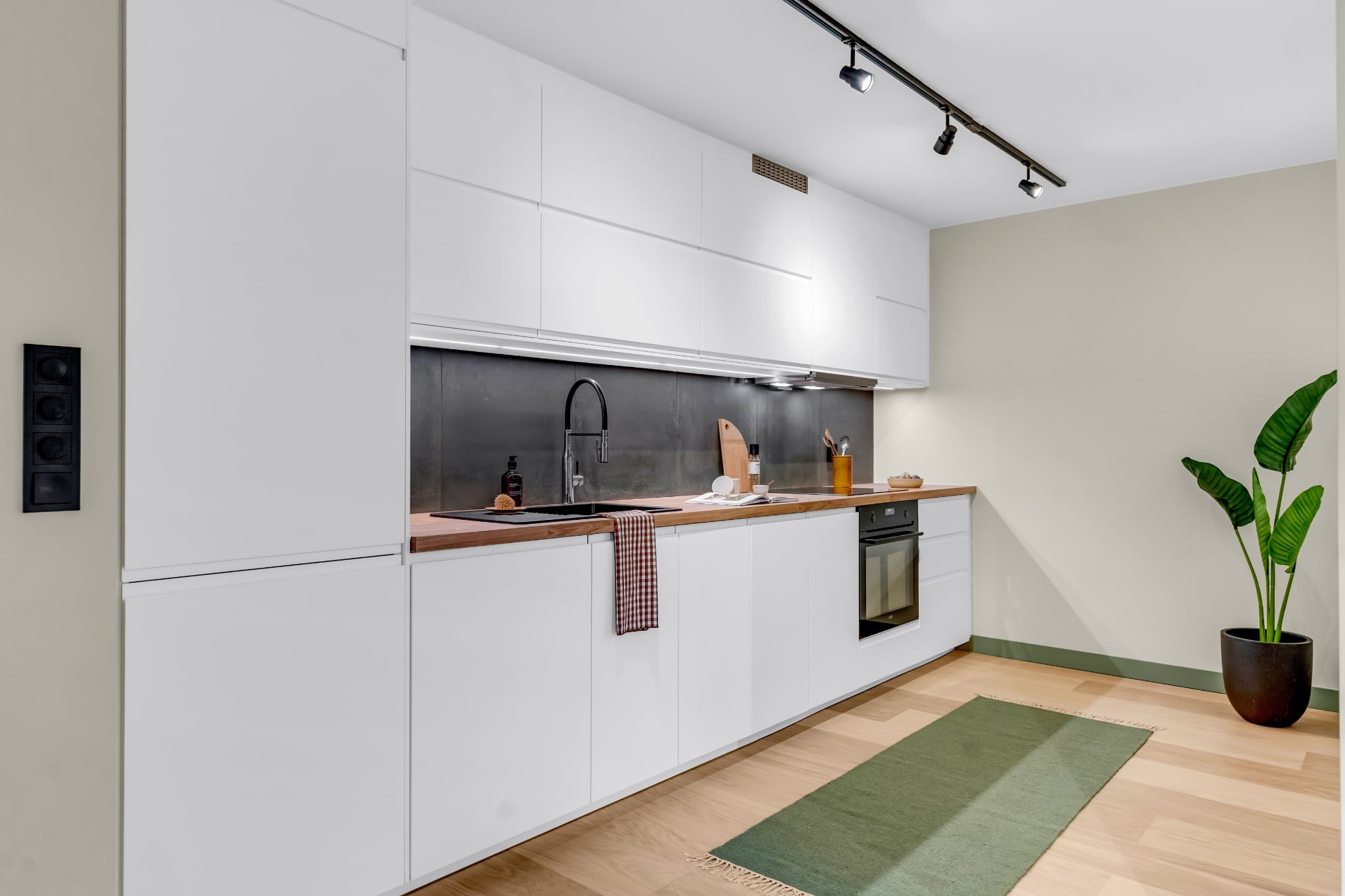 Nowoczesna kuchnia z białymi szafkami, drewnianym blatem i czarnym panelem nad zlewem. Oświetlenie punktowe, roślina doniczkowa, dywanik. Minimalistyczny design.