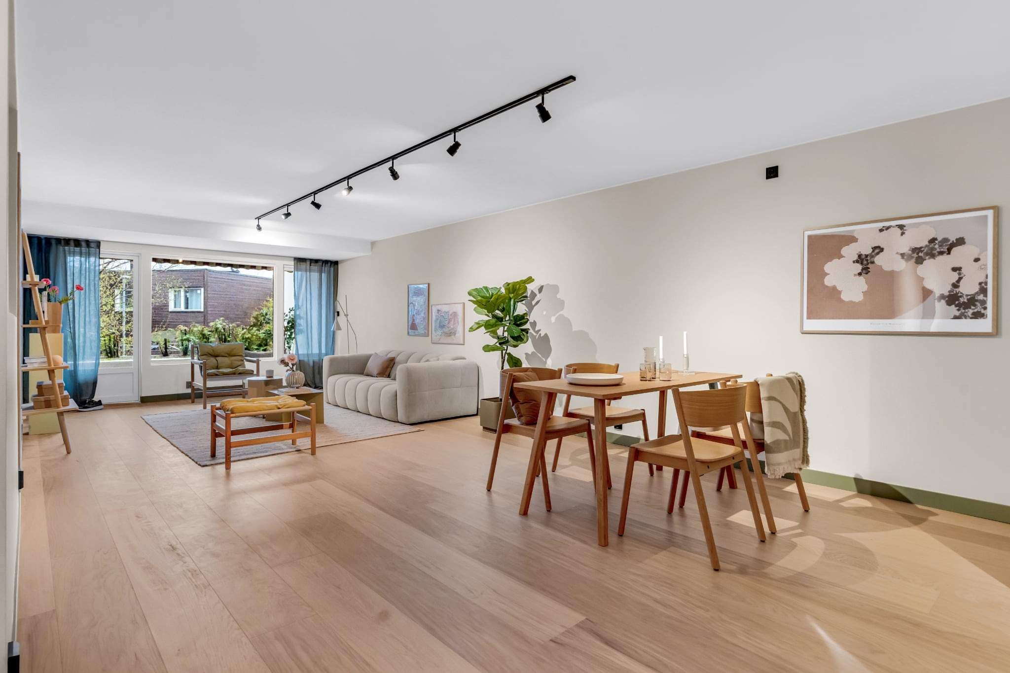 Jasne, przestronne wnętrze z drewnianą podłogą i nowoczesnymi meblami. Widoczny stół z krzesłami, sofa i dekoracje. Całość utrzymana w neutralnej kolorystyce z akcentami zieleni.