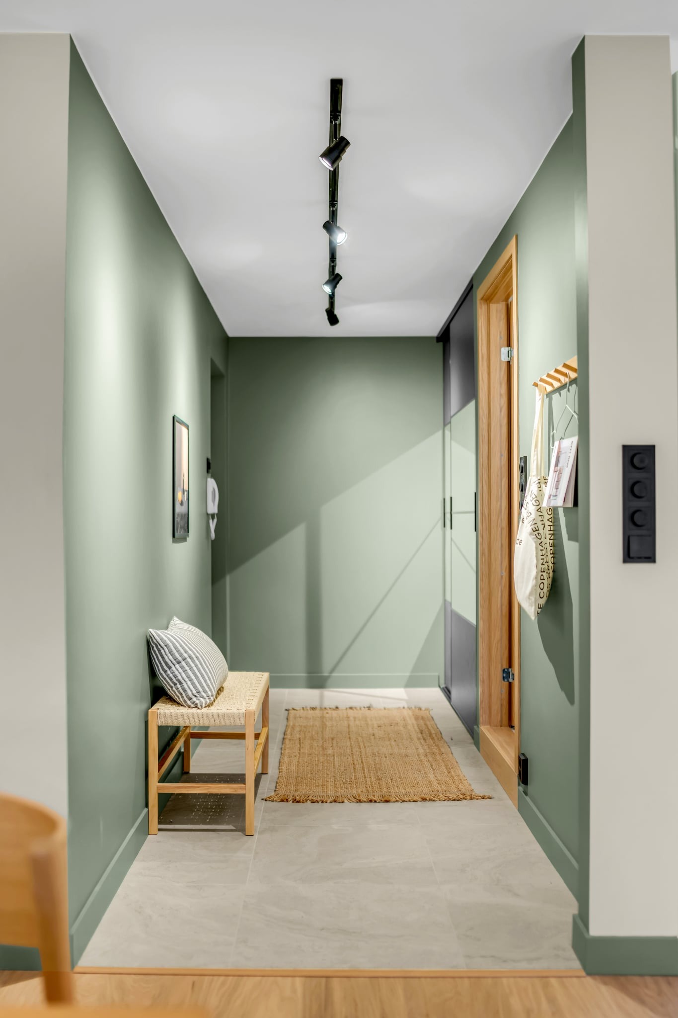 Stylowy korytarz w odcieniach zieleni z drewnianą ławką, jutowym dywanikiem i oświetleniem punktowym. Widoczne drzwi przesuwne i dekoracyjne akcenty.