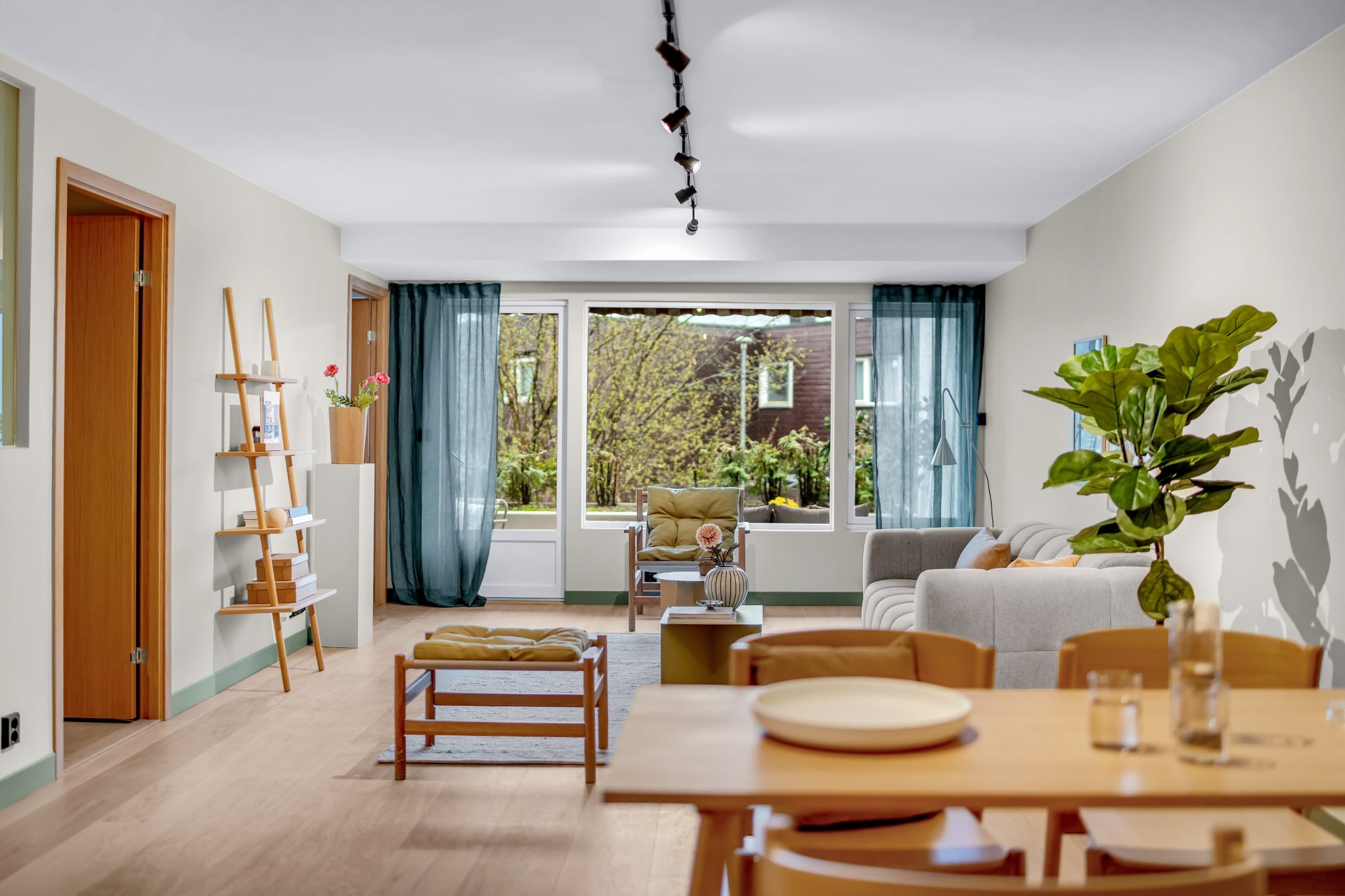 Jasne, przestronne wnętrze salonu z drewnianymi meblami, zielonymi akcentami i dużym oknem z widokiem na zieleń. Minimalistyczny design z roślinami doniczkowymi.