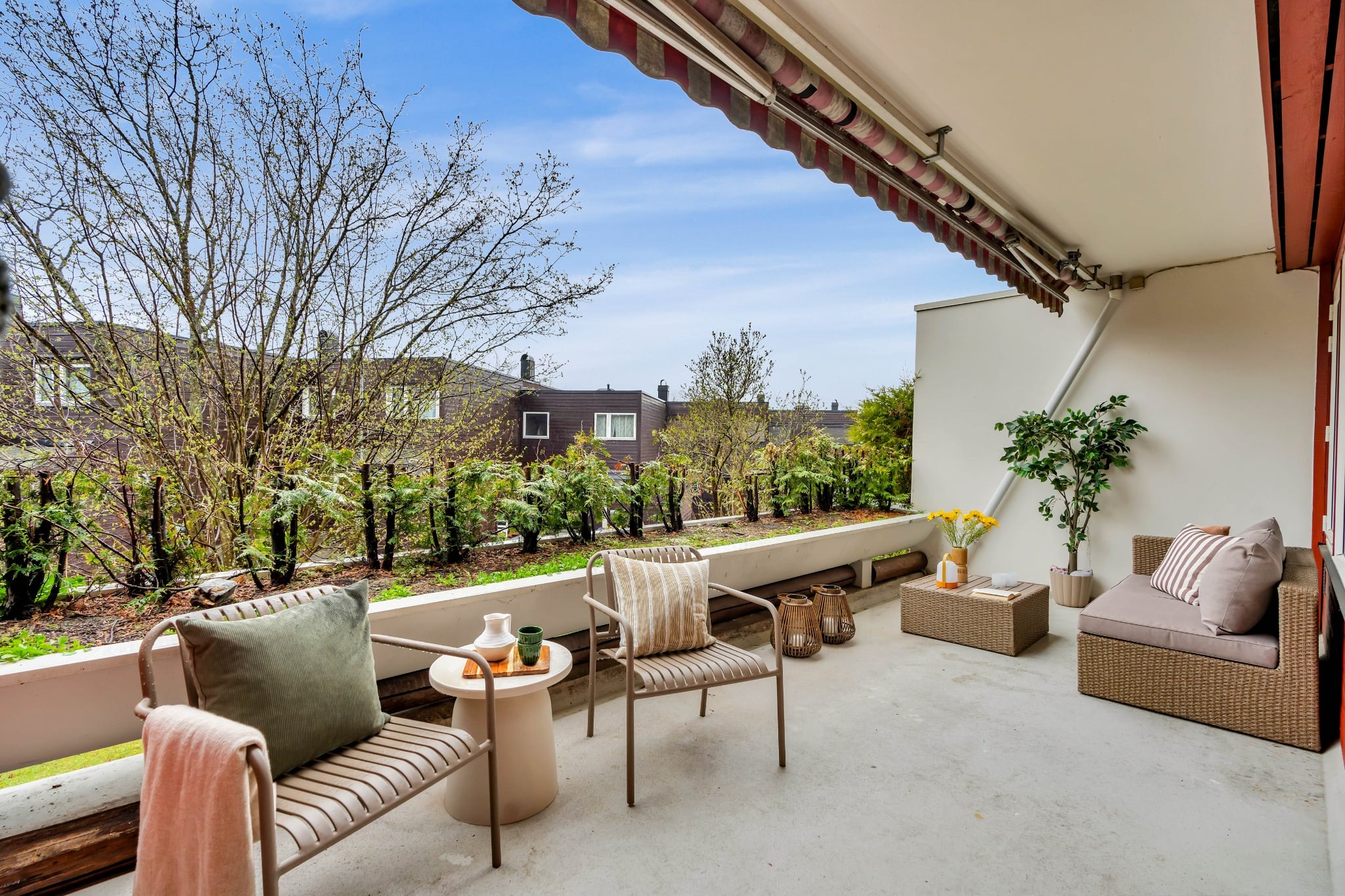 Umeblowany balkon z markizą i widokiem na zieleń. Dwa krzesła, stolik i sofa tworzą przytulną przestrzeń. Rośliny wzdłuż balustrady dodają naturalnego akcentu.