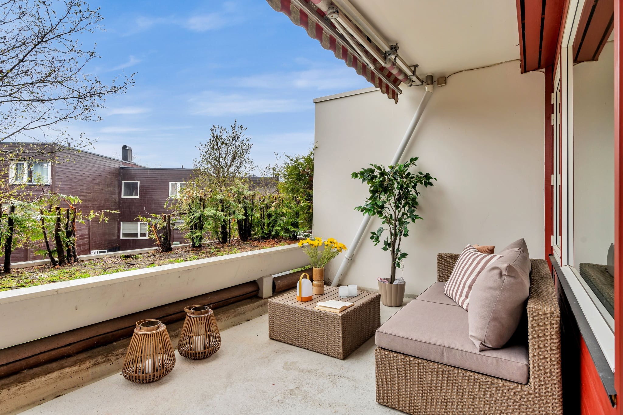 Zadaszony balkon z meblami ogrodowymi, roślinami w donicach i widokiem na zielony dach budynku. Stolik kawowy, lampiony i sofa tworzą przytulną przestrzeń.