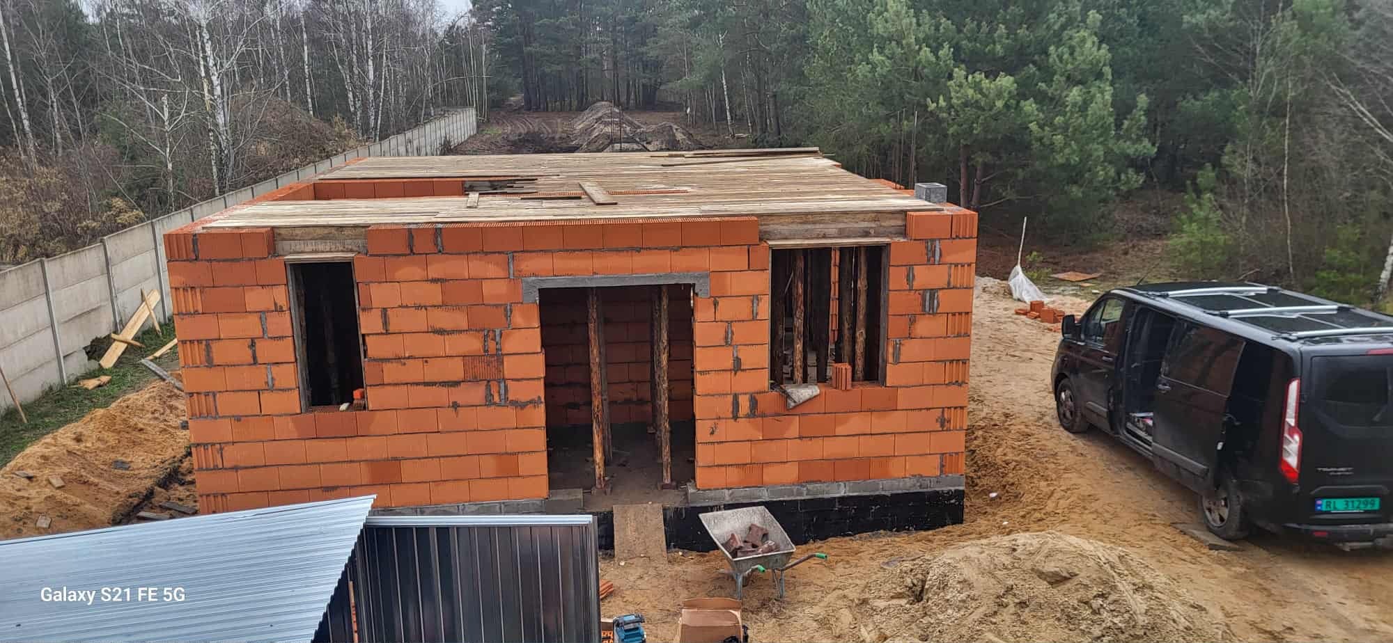 Budowa domu z czerwonej cegły, widoczne otwory okienne i drzwiowe, częściowo zadaszony, taczka z cegłami obok, czarny van w tle, teren budowy.