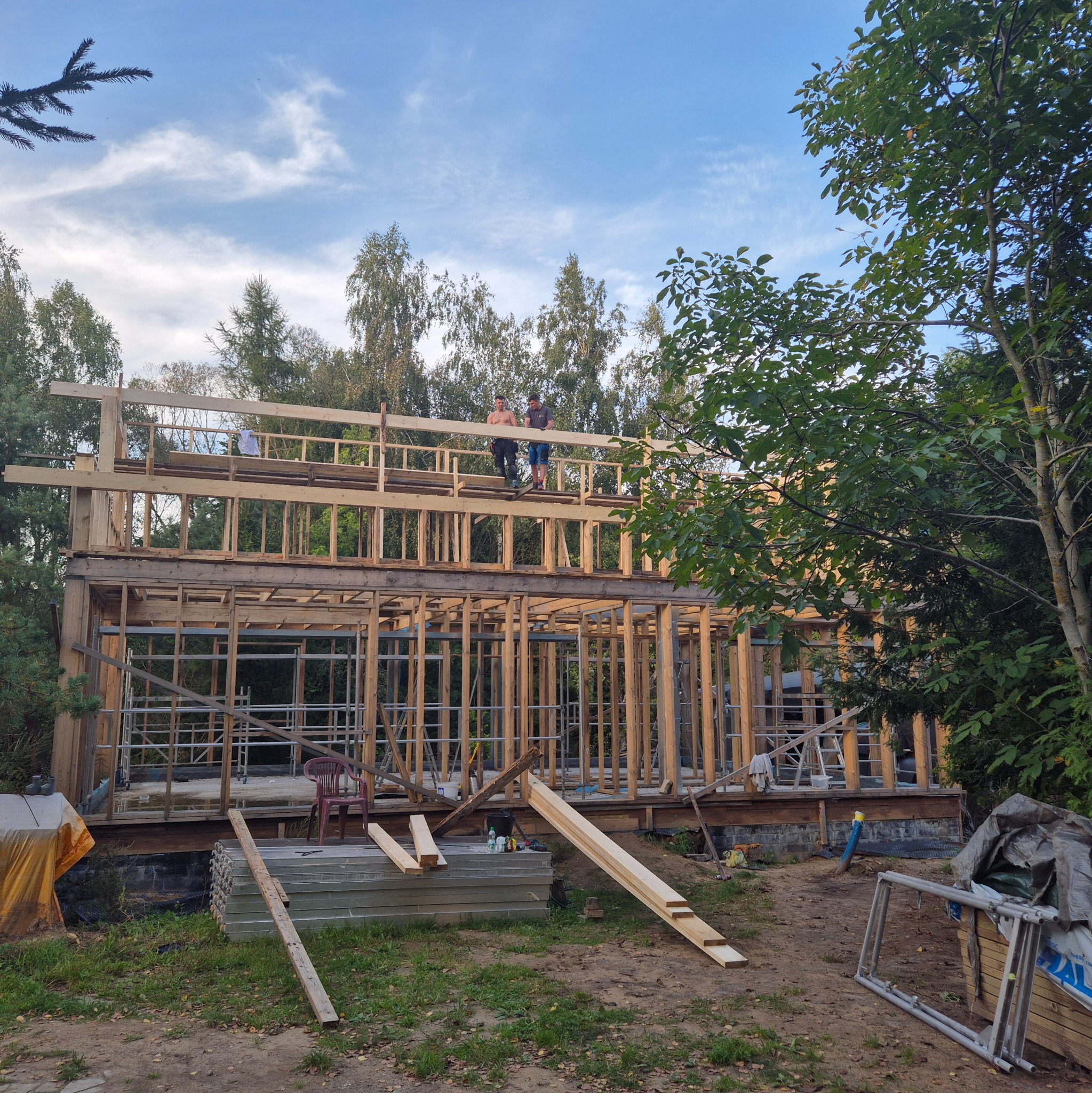 Budowa domu szkieletowego w lesie: dwukondygnacyjna konstrukcja z drewna, dwóch mężczyzn na górze, materiały budowlane na ziemi, drzewa w tle.