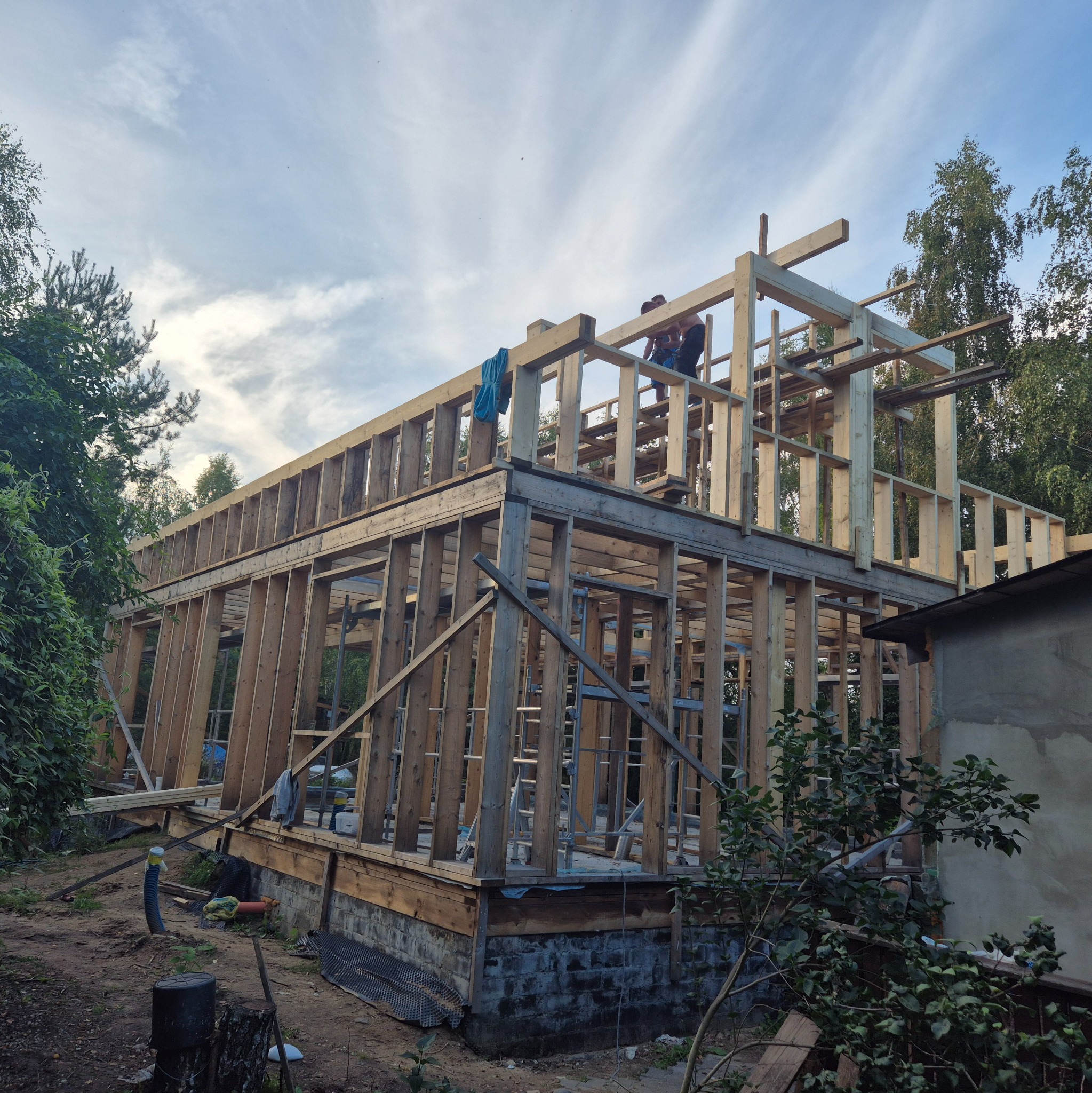Konstrukcja domu szkieletowego w Wyszkowie w trakcie budowy. Widoczne drewniane elementy konstrukcyjne i pracownicy na górze. Tło stanowią drzewa i niebo.