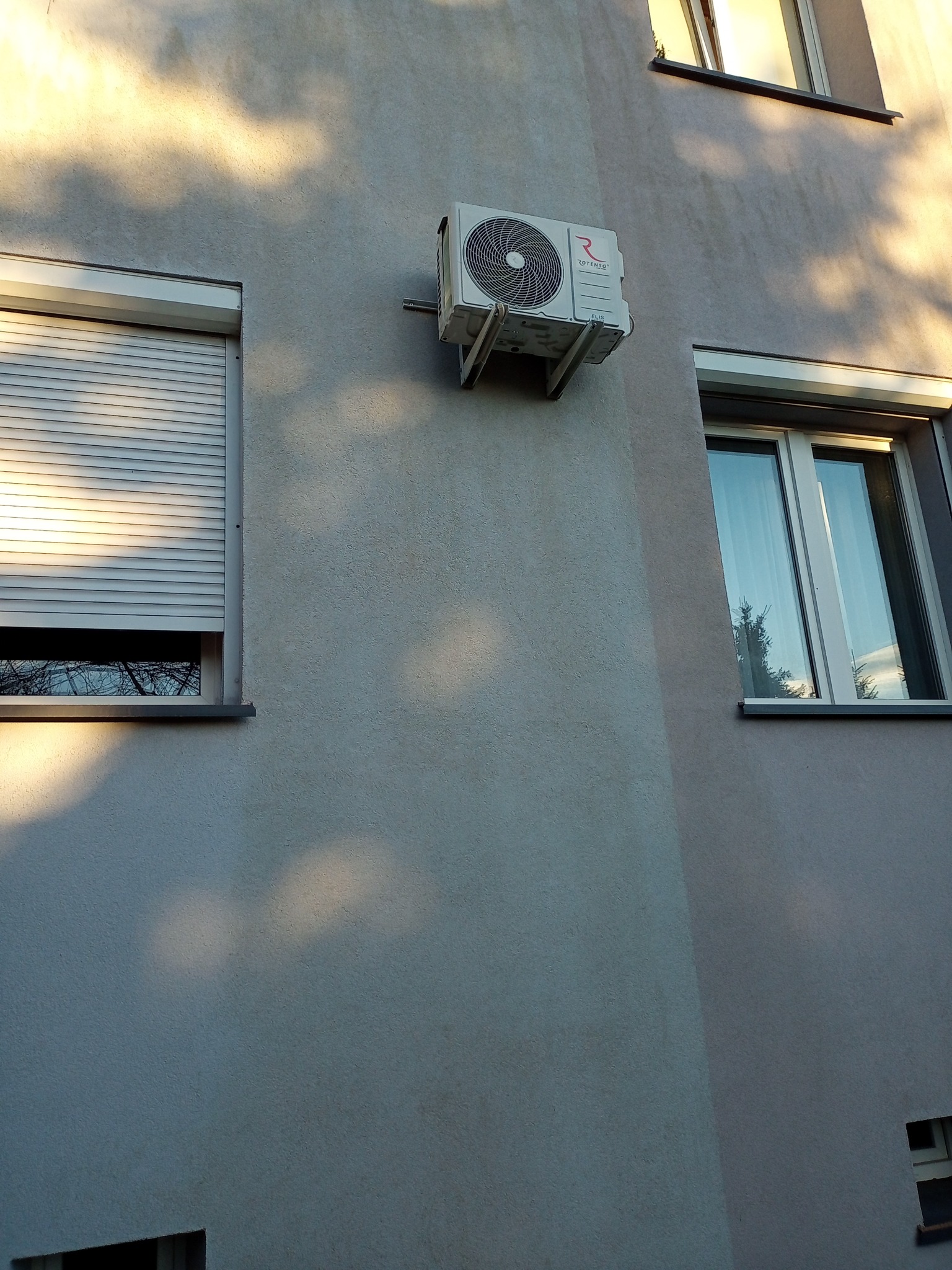 Zamontowana klimatyzacja Rotenso na elewacji budynku mieszkalnego we Wrocławiu, widoczna z boku. W tle okna z roletami i odbicie drzew w szybie.