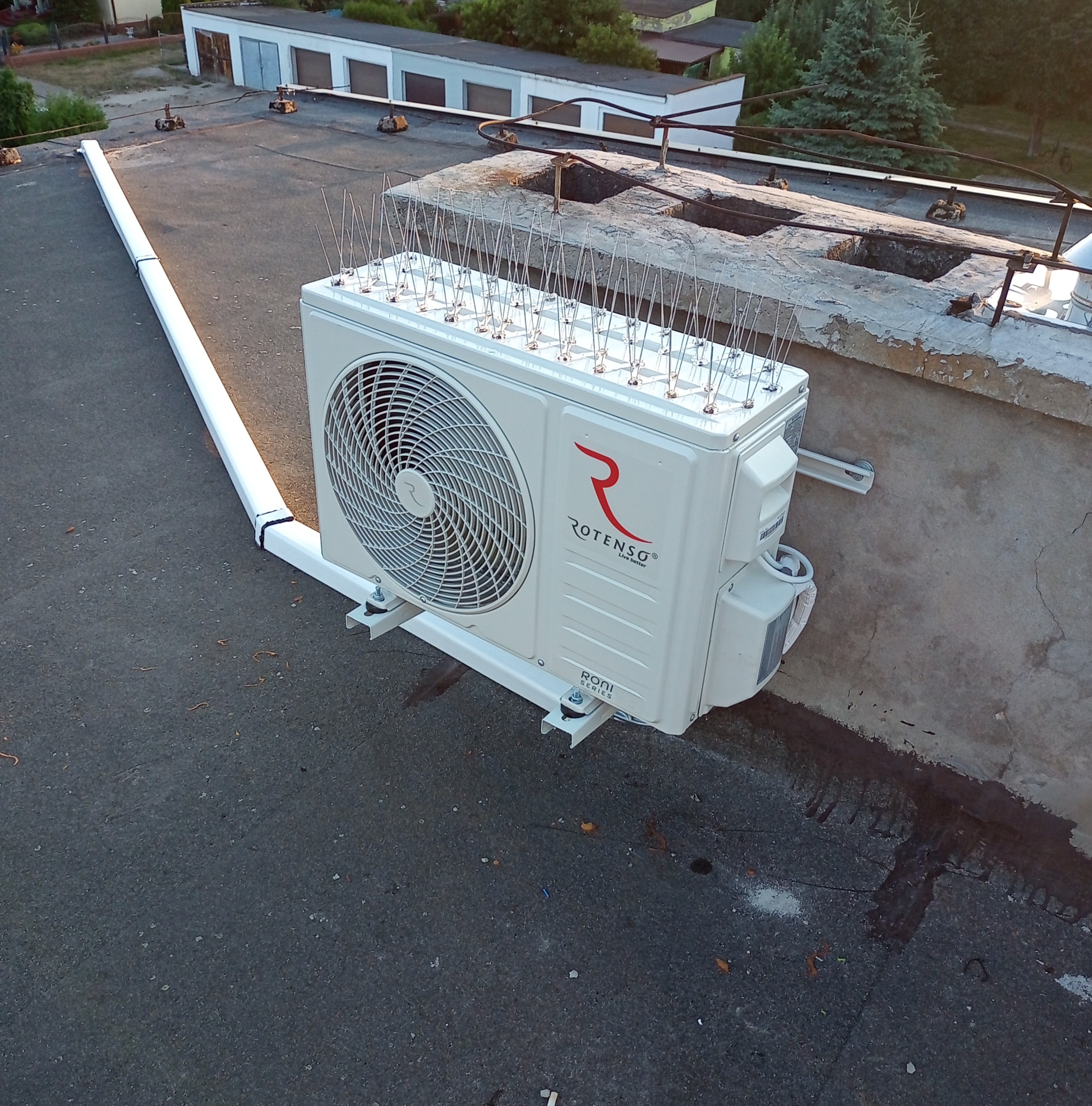 Biały klimatyzator Rotenso, zabezpieczony kolcami przeciw ptakom, zamontowany na dachu budynku. Widoczne elementy instalacji i tło z budynkami.