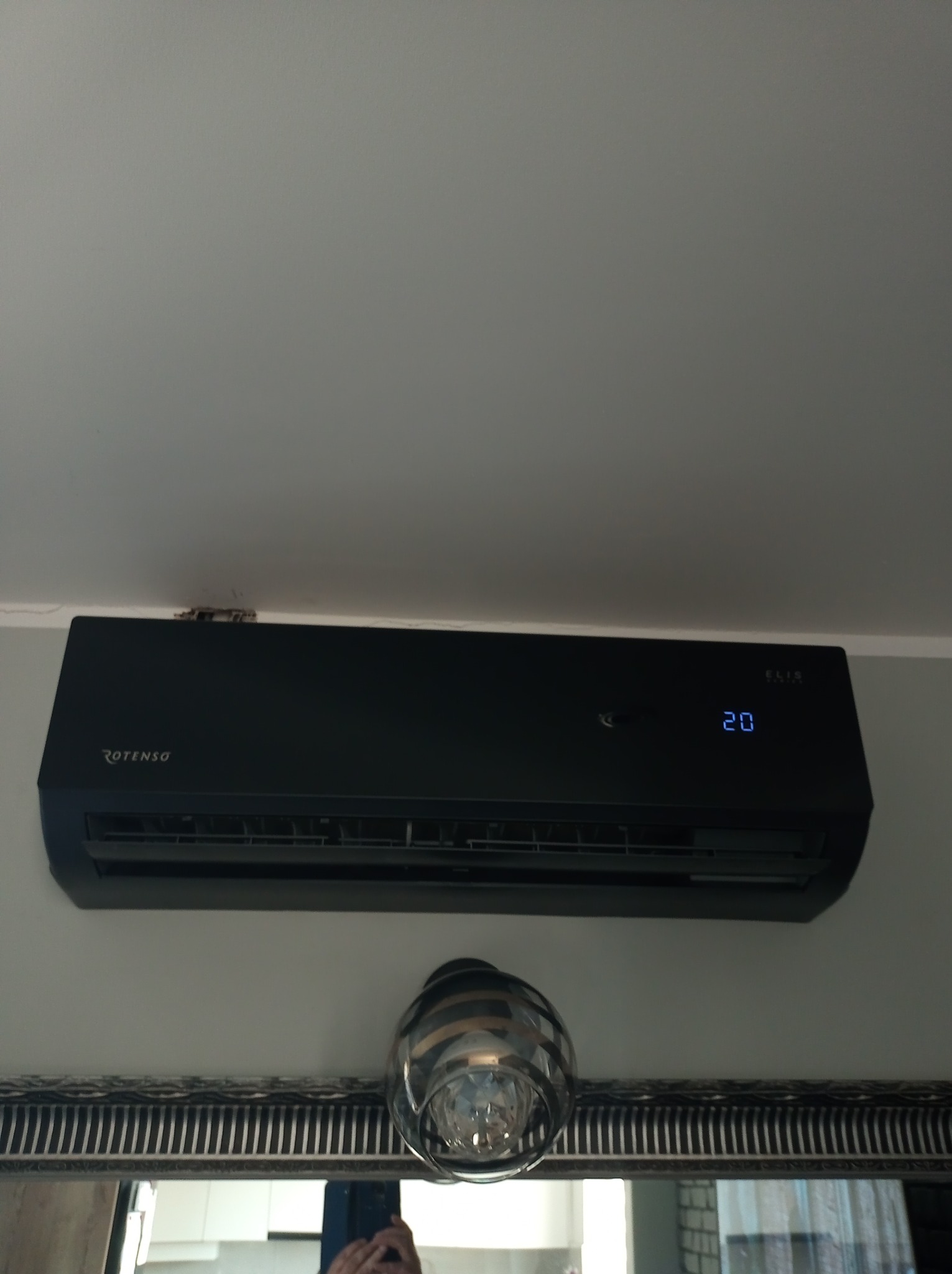 Czarny klimatyzator Rotenso Elixir zamontowany na ścianie nad lustrem z dekoracyjną lampą. Widoczny wyświetlacz temperatury.