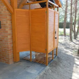 Drewniana kabina prysznicowa na zewnątrz z lamelowymi drzwiami, widoczna instalacja solarna na dachu, położona na płytkach i piasku, obok ściana z cegły.