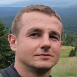 Portret mężczyzny na tle górskiego krajobrazu. Krótkie włosy, lekki zarost, stonowana kolorystyka. Widoczne drzewa i wzgórza w oddali. Ujęcie od przodu, naturalne światło.