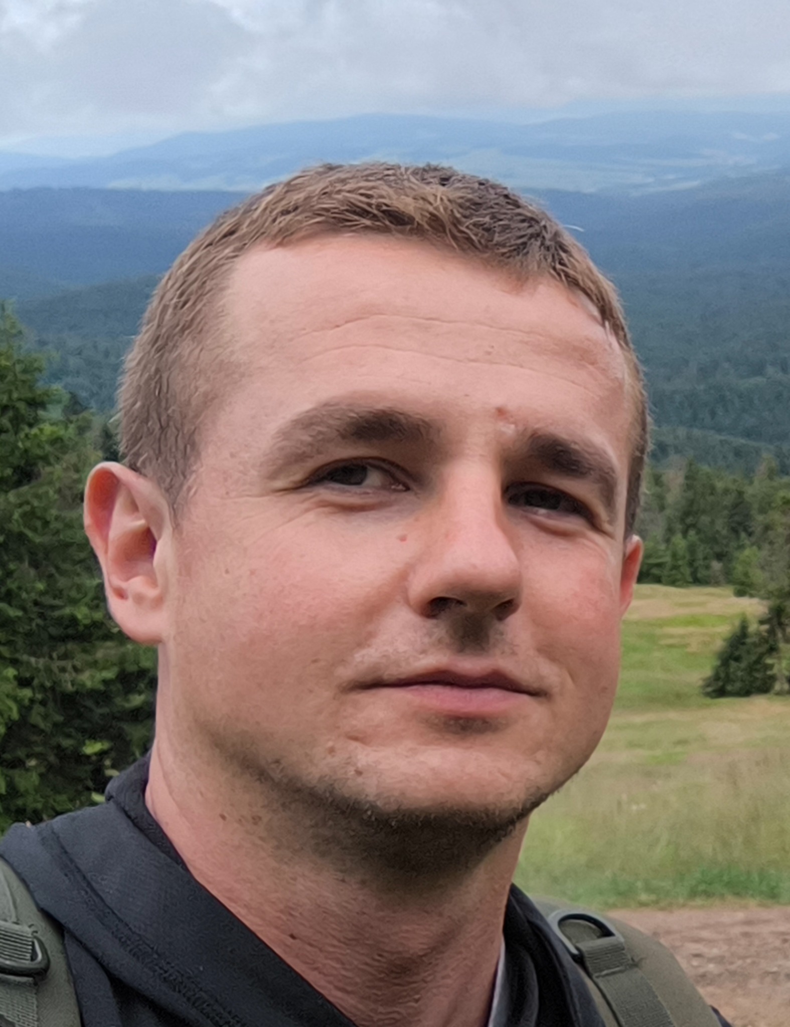 Portret mężczyzny na tle górskiego krajobrazu. Krótkie włosy, lekki zarost, stonowana kolorystyka. Widoczne drzewa i wzgórza w oddali. Ujęcie od przodu, naturalne światło.