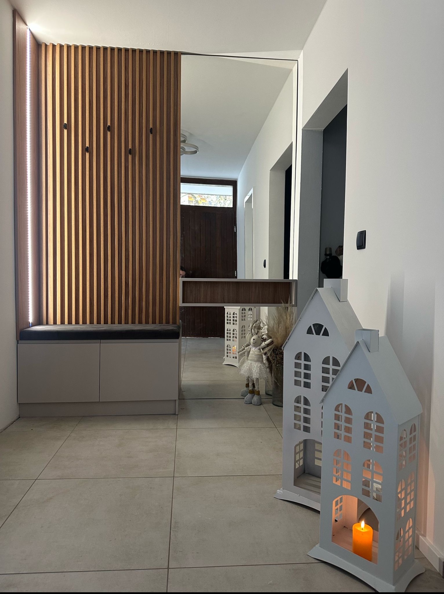 Nowoczesny przedpokój z drewnianym panelem ściennym, siedziskiem z szarej tapicerki i dekoracyjnymi lampionami w kształcie domków. Lustro optycznie powiększa przestrzeń.