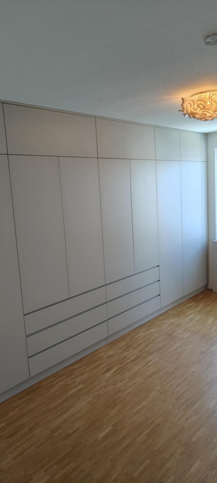 Jasnoszara szafa wnękowa z szufladami, zajmująca całą ścianę, z drewnianą podłogą i ozdobną lampą sufitową. Minimalistyczny design.